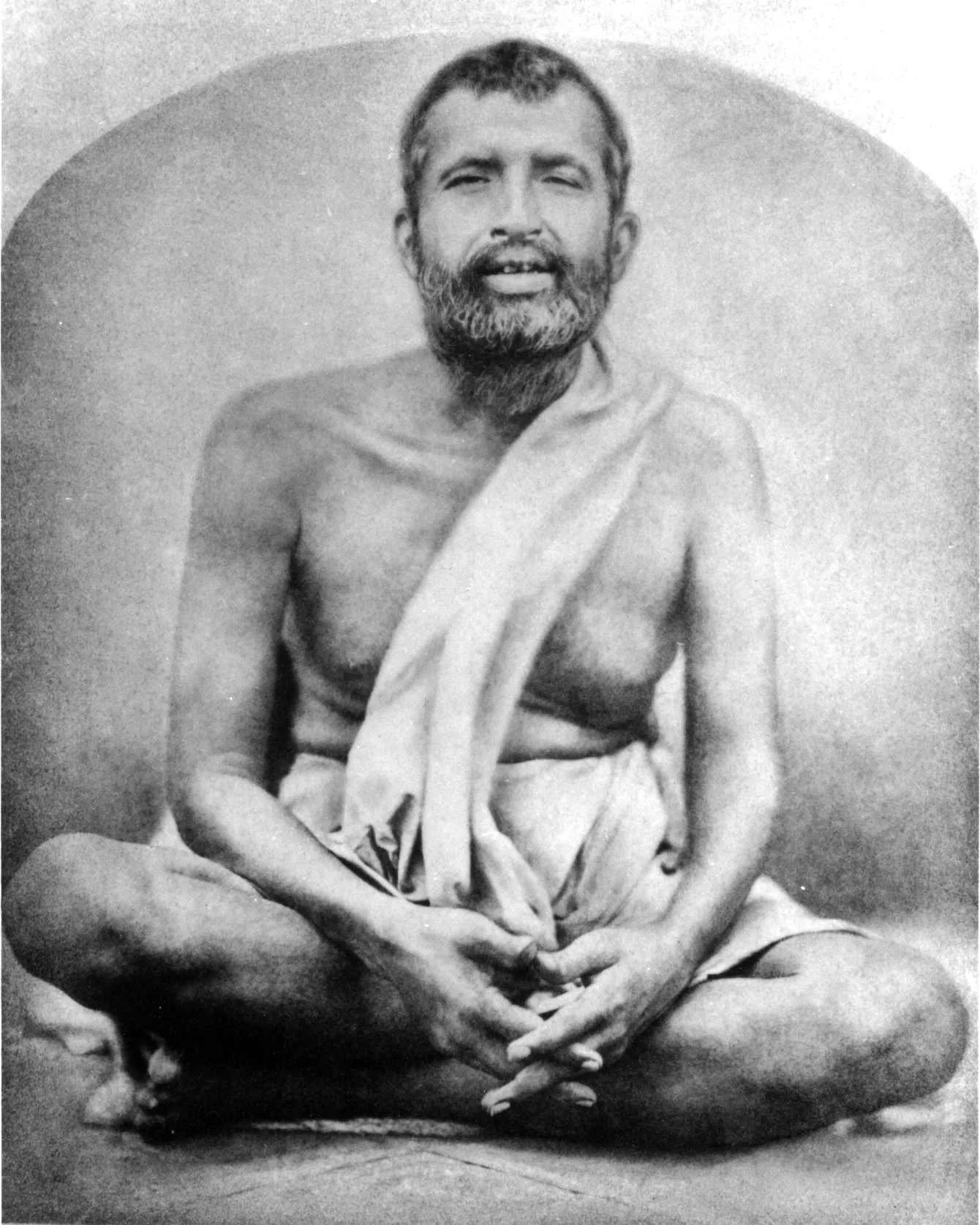 A black and white photograph of Ramakrishna, sitting cross-legged, wearing traditional clothing draped over the shoulder, and seated against a plain background.