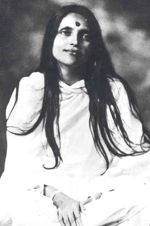 Black and white photograph of Anandamayi Ma wearing traditional Indian attire, sitting with her hand resting on her lap, looking at the camera.
