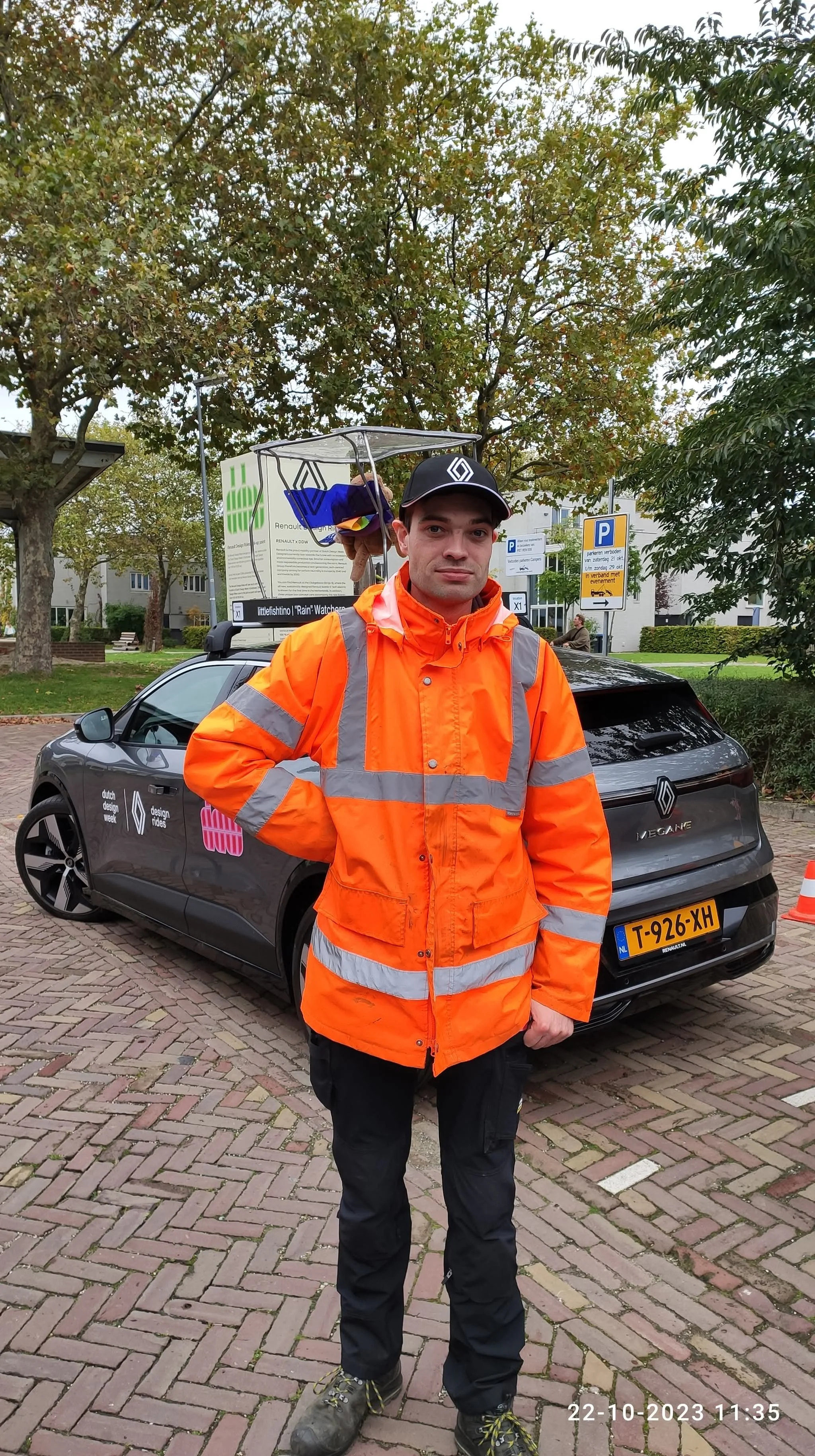 A person in an orange safety jacket and black cap stands in front of a car with Dutch design branding, near a parking sign and trees.