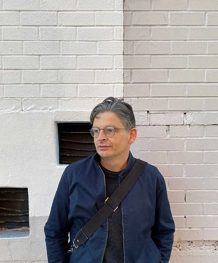 Person with glasses wearing a navy jacket and a crossbody bag, standing against a white brick wall with small windows.