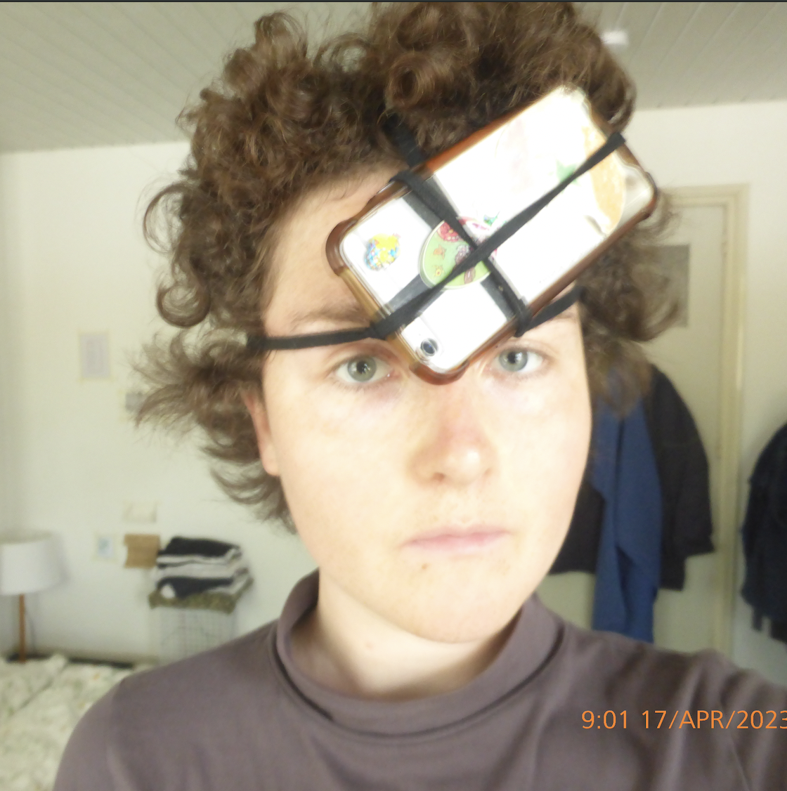 Person with a phone strapped to their forehead using black bands, in a bedroom setting with clothing and lamp in the background.