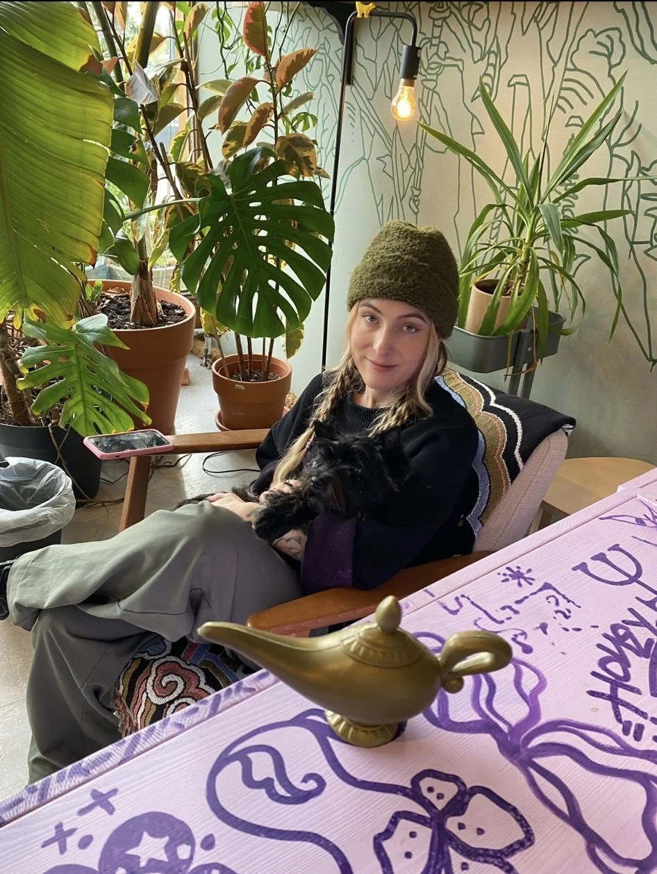 Person wearing a green beanie sitting in a cozy, plant-filled room, holding a small black dog. A decorative table with a purple design and a golden genie lamp is in the foreground. Potted plants and ambient lighting create a warm atmosphere.
