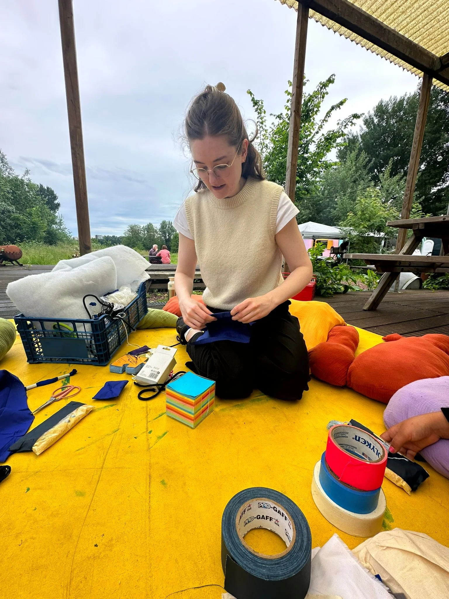 A person sitting on a yellow mat outdoors, surrounded by crafting materials like colored tape, fabric, and scissors. They are wearing glasses and a beige sleeveless top over a white shirt. Trees and greenery are visible in the background.