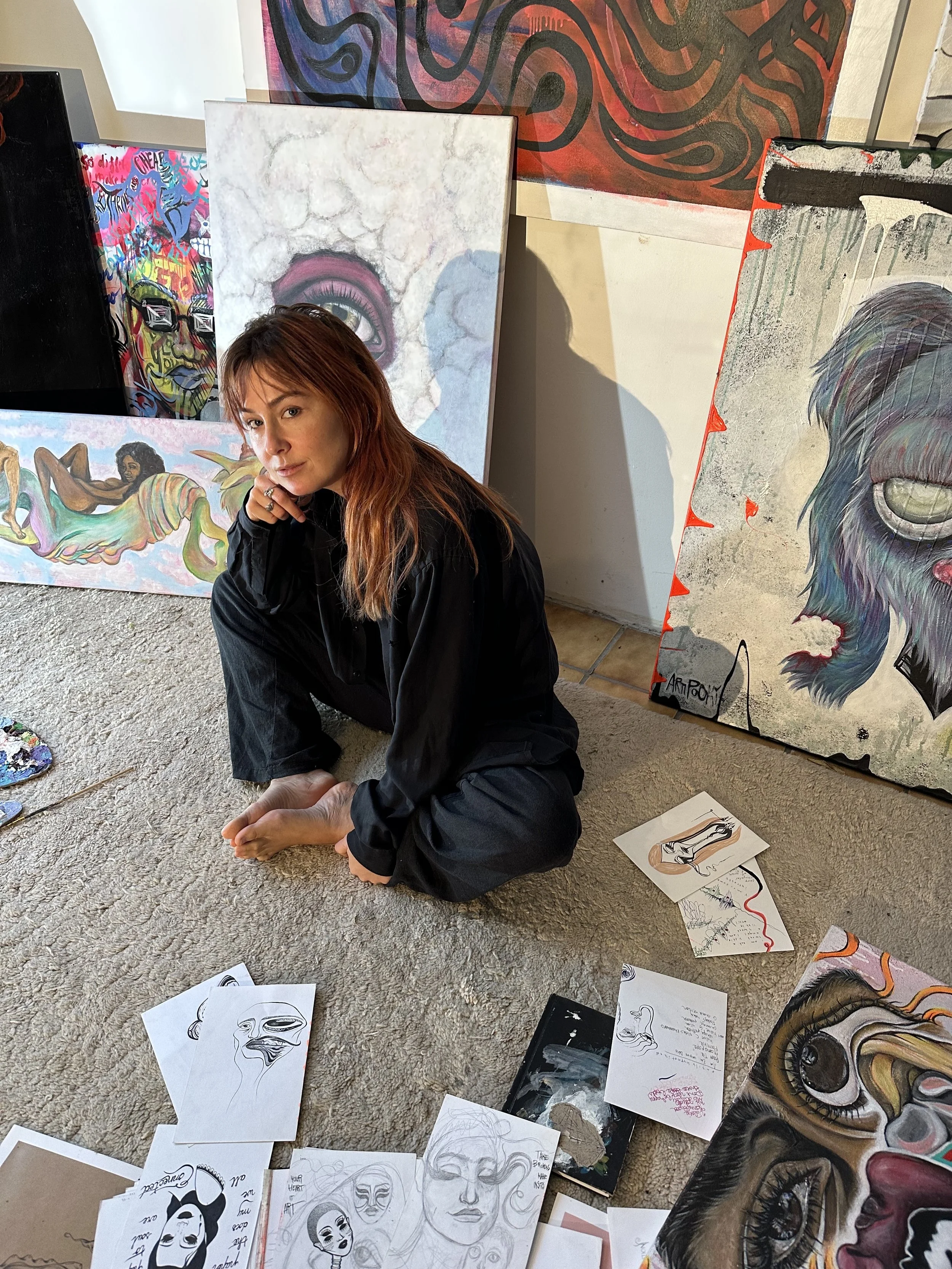Artist surrounded by various artworks, including paintings on canvases and sketches on paper, seated on a textured carpet in a creative studio space.