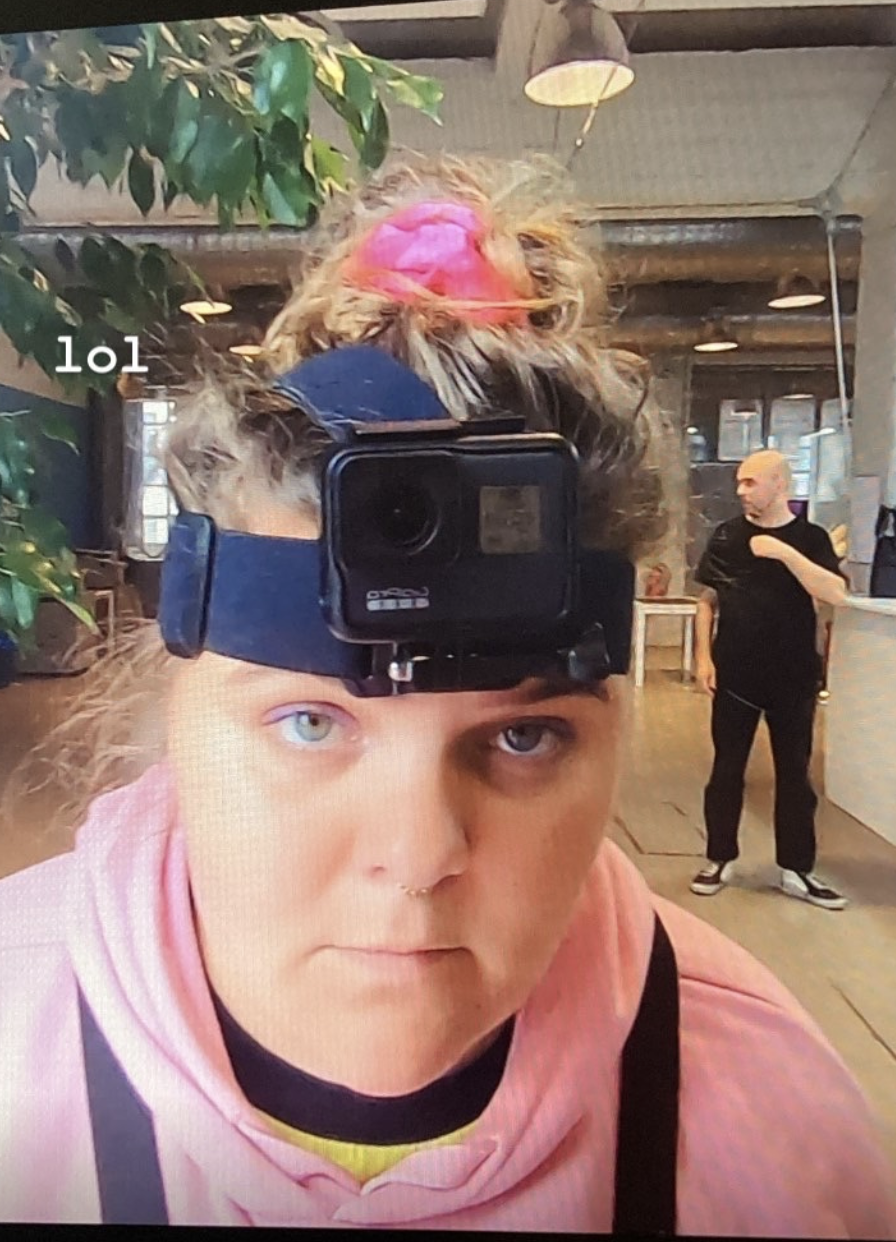 Person wearing a head-mounted camera in an indoor setting with another person in the background.