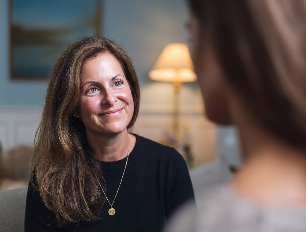 Phoebe Sade, A woman with long brown hair and a black shirt smiling during a conversation, sitting in a cozy room with a lamp in the background.
