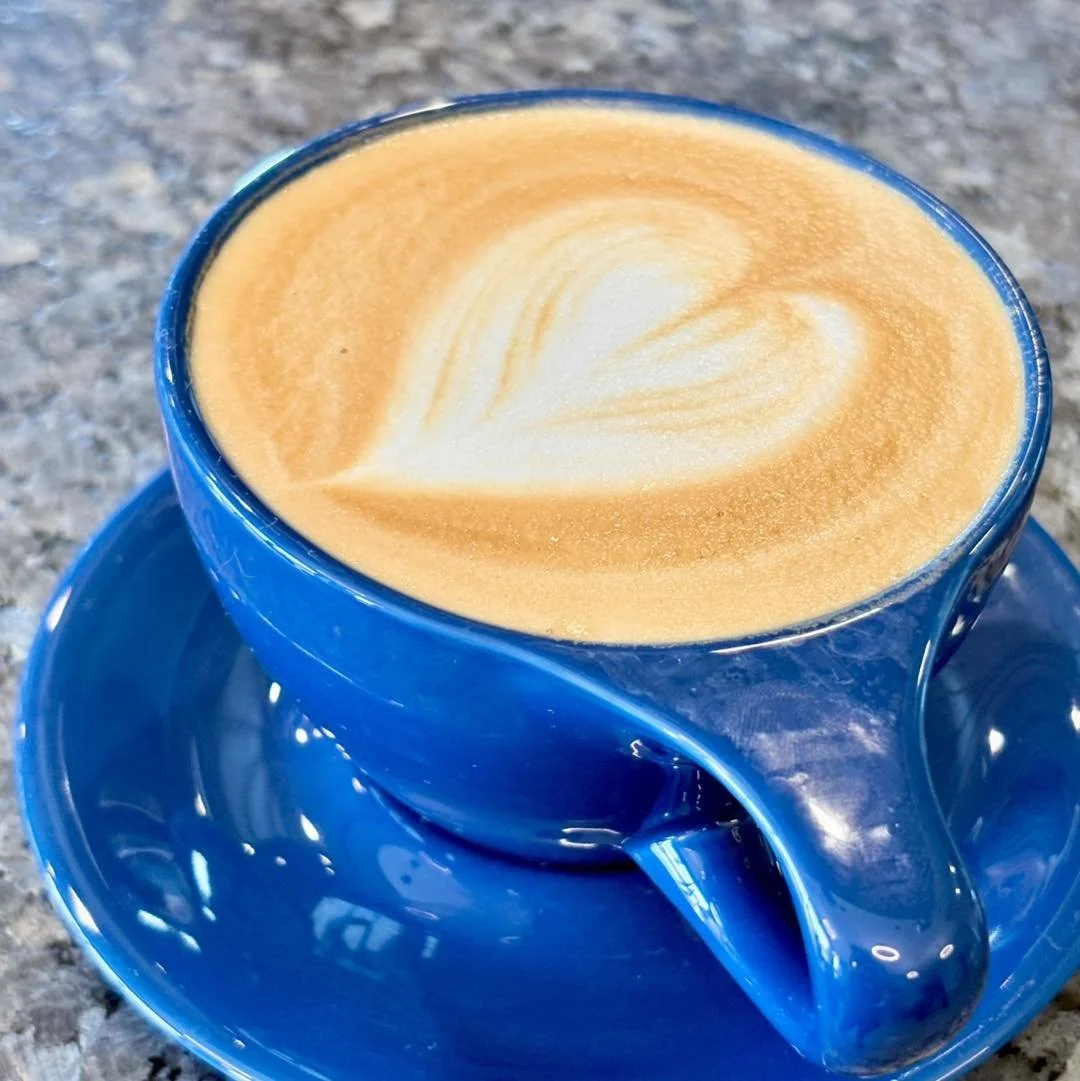 A blue ceramic cup filled with cappuccino topped with foam latte art in the shape of a heart, placed on a matching blue saucer on a granite countertop.