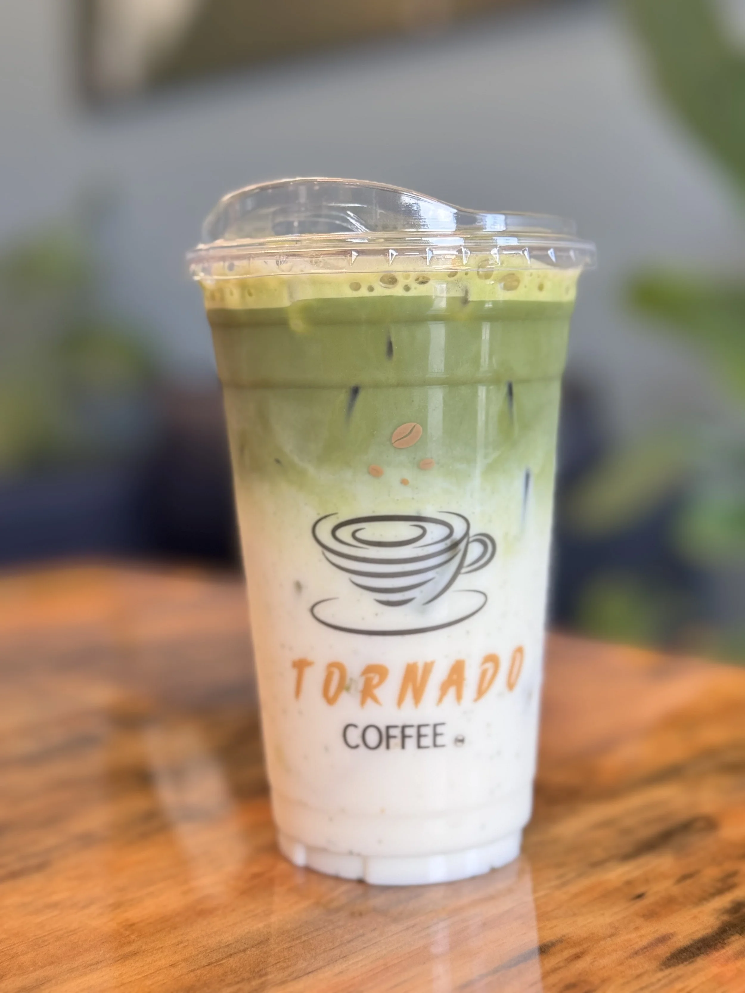 Close-up of a plastic cup with green matcha latte topped with foam, sitting on a wooden table.