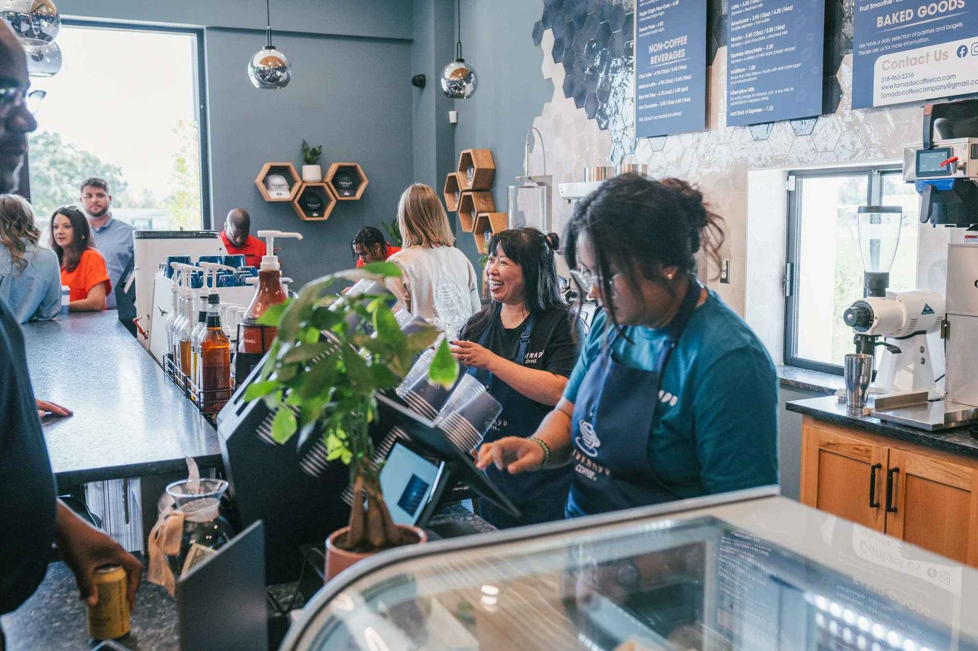 Inside a coffee shop with customers and staff at the counter, menu boards on the wall, and a large window letting in natural light.