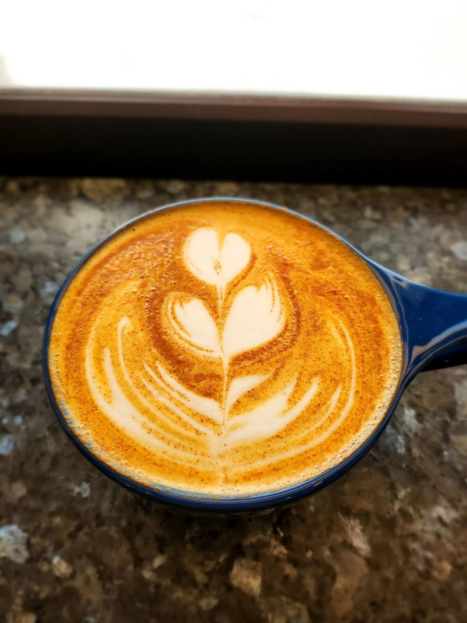 A close-up view of a coffee cup with latte art in the shape of a tulip design on top, placed on a speckled brown countertop.