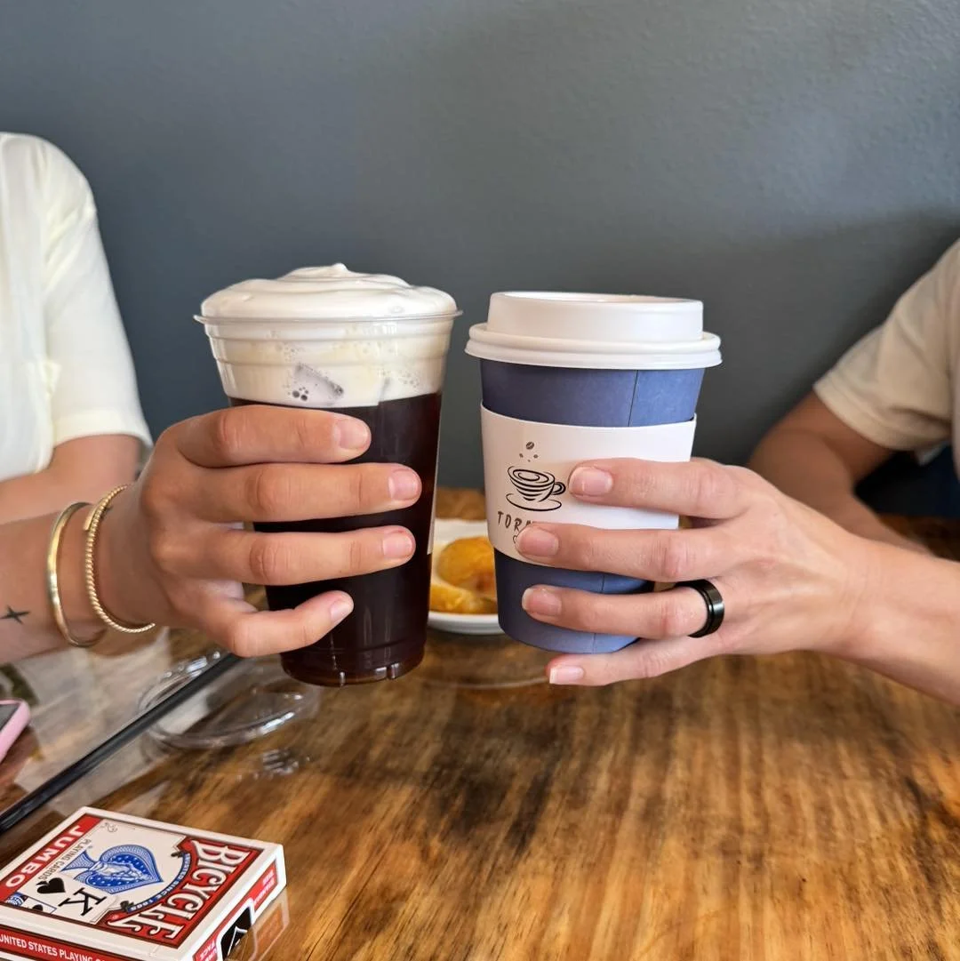 Two people clinking coffee cups over a wooden table, with a plate of snacks and a deck of cards visible in the background.