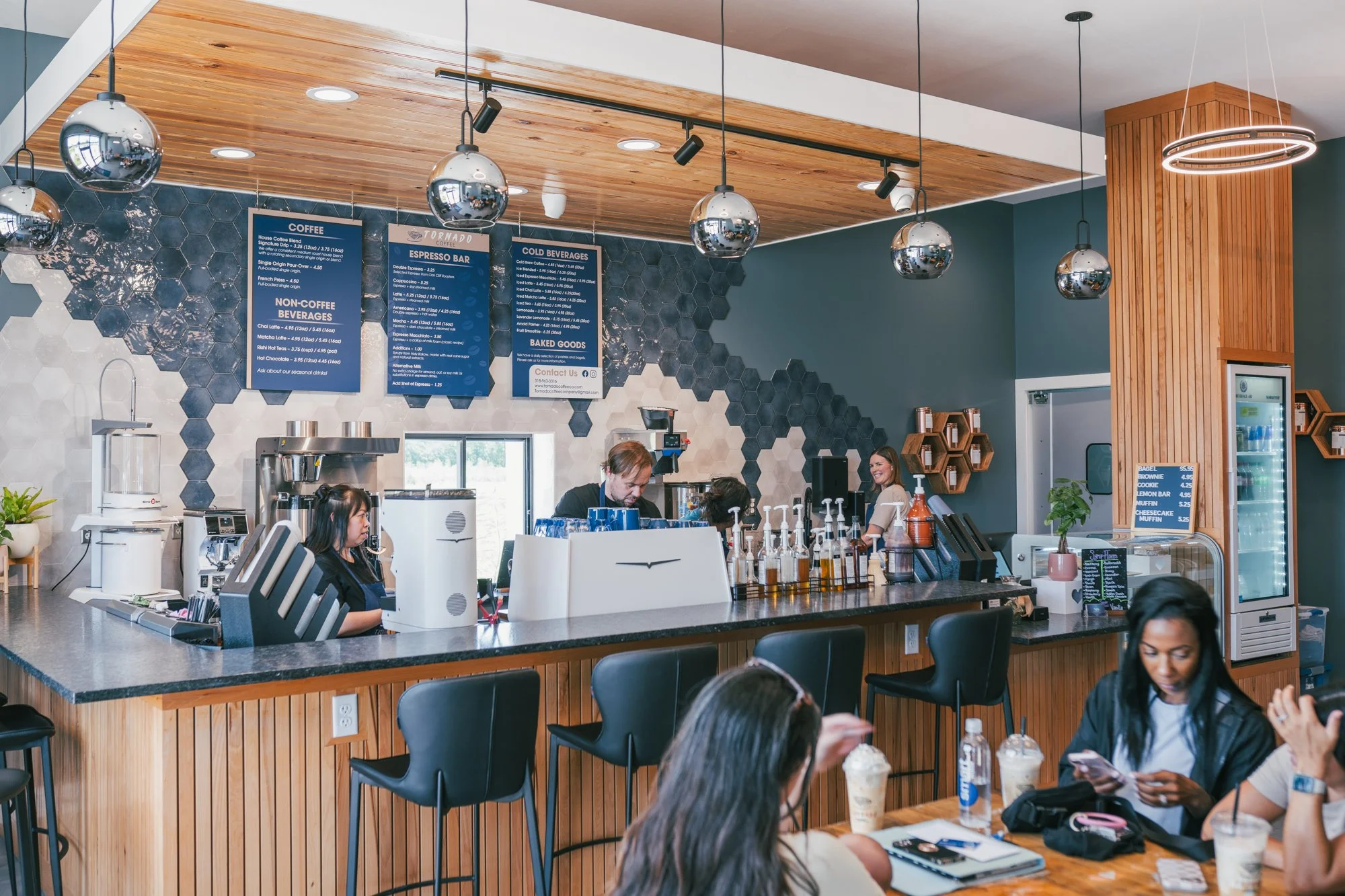 Interior of a modern coffee shop with a counter where baristas are preparing beverages, menu boards hanging from the light wood ceiling, and customers seated at tables enjoying drinks and chatting.