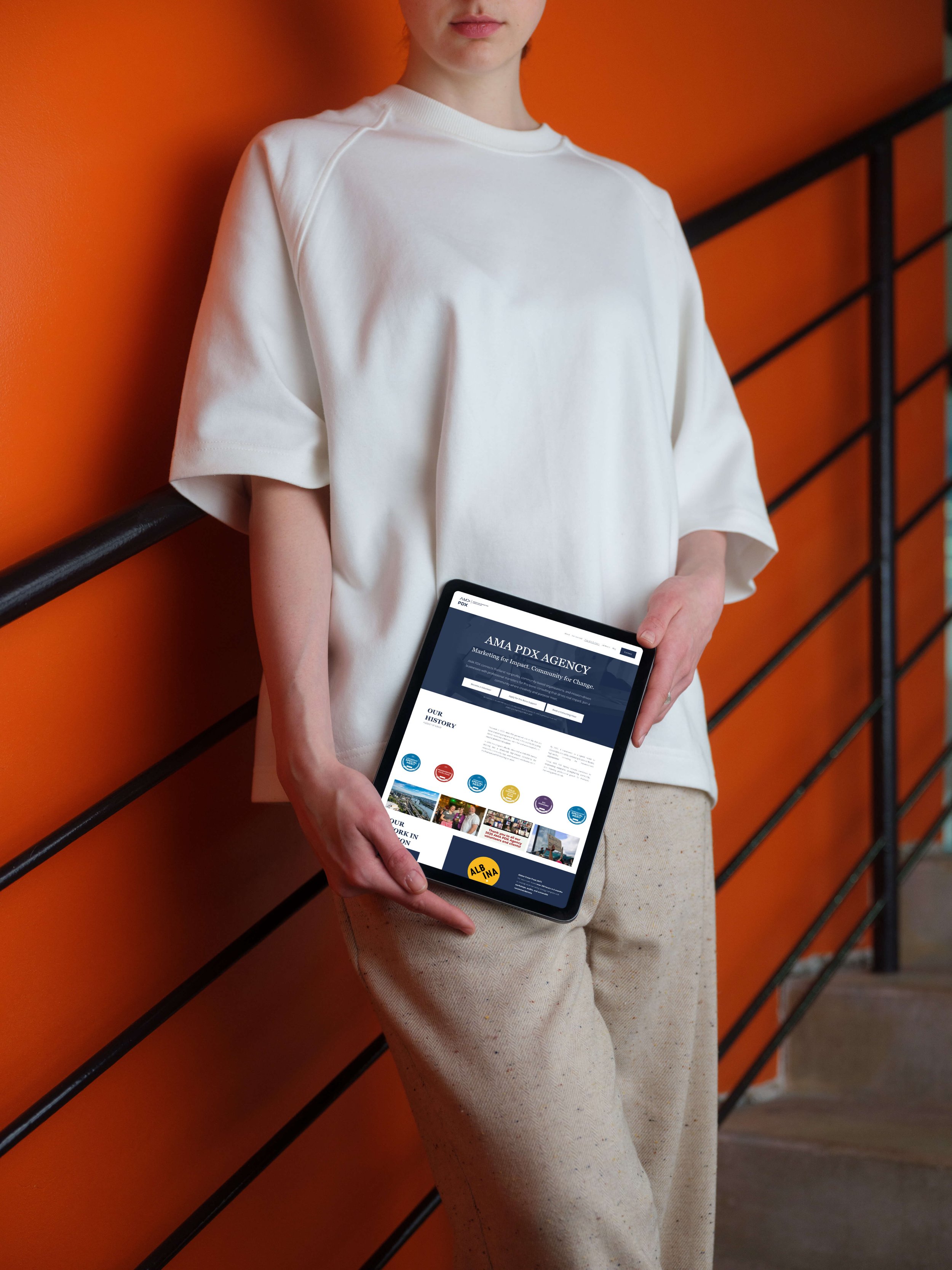 Person holding a tablet with a website displayed, standing against an orange wall and black railing.