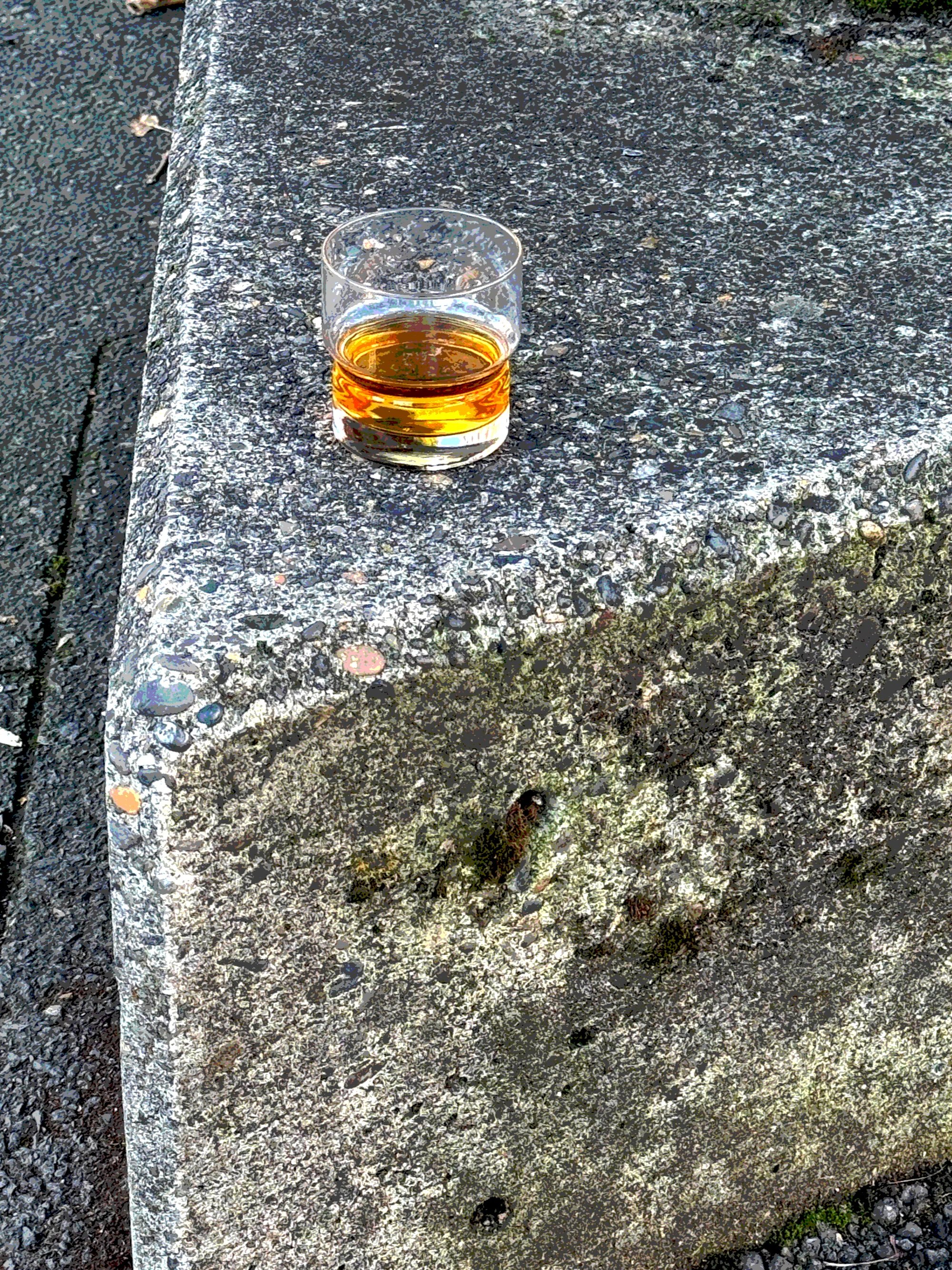 A glass of amber-colored liquid, possibly whiskey or bourbon, placed on a rough concrete surface outdoors.