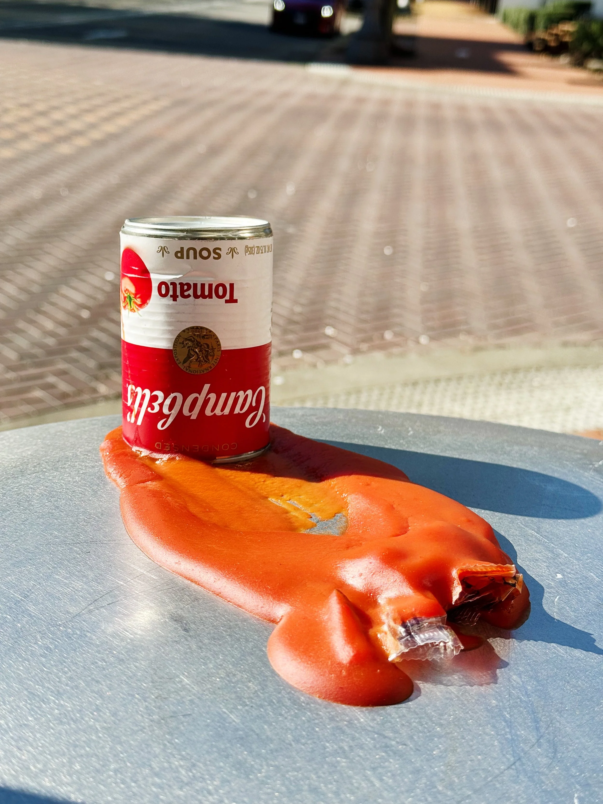 A can of tomato soup placed upside-down on a melted mess of ketchup on a metal surface with an outdoor background.