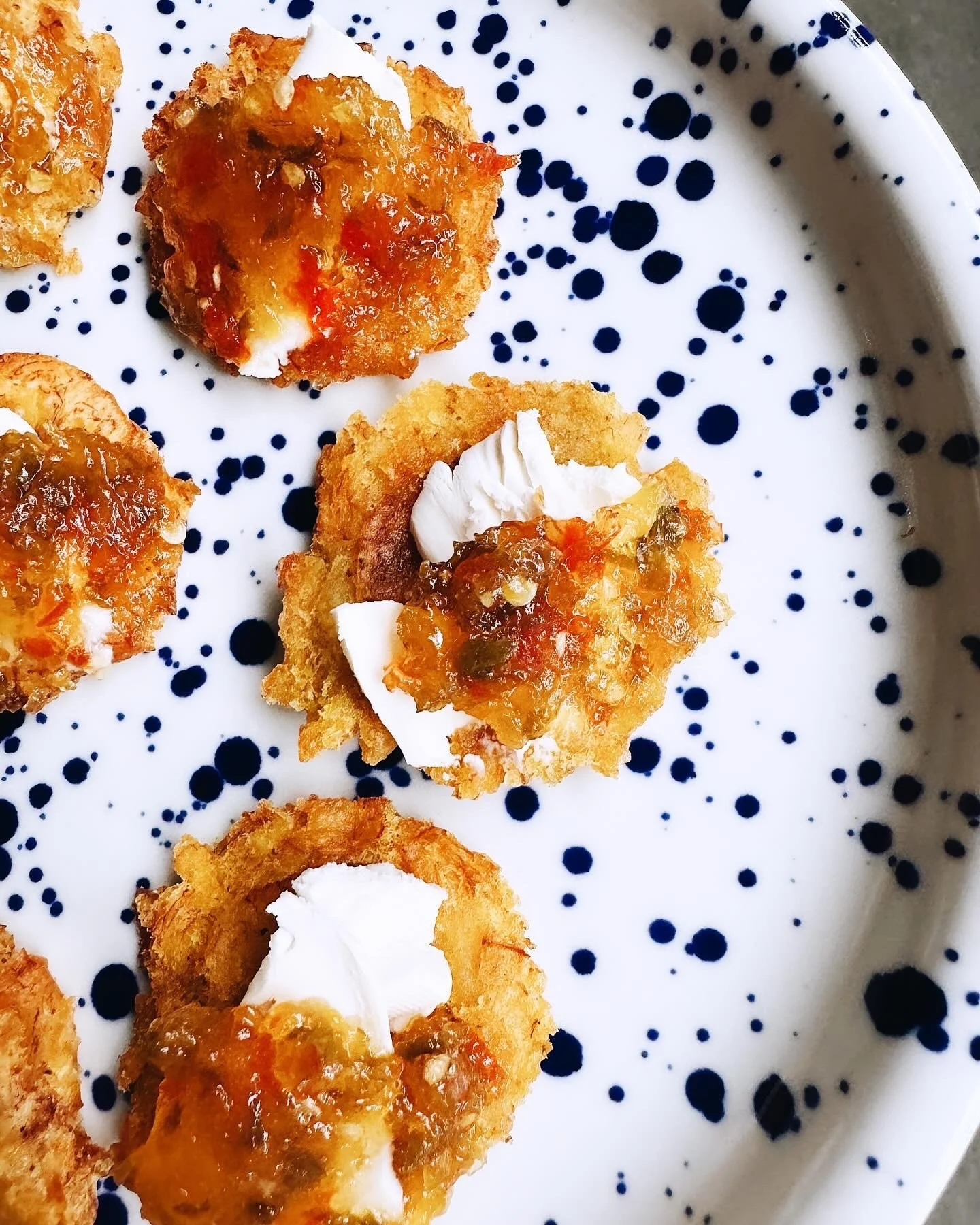 Close-up of five breaded and fried appetizers topped with a dollop of white sauce and topped with orange jam, served on a white plate with navy blue dotted pattern.
