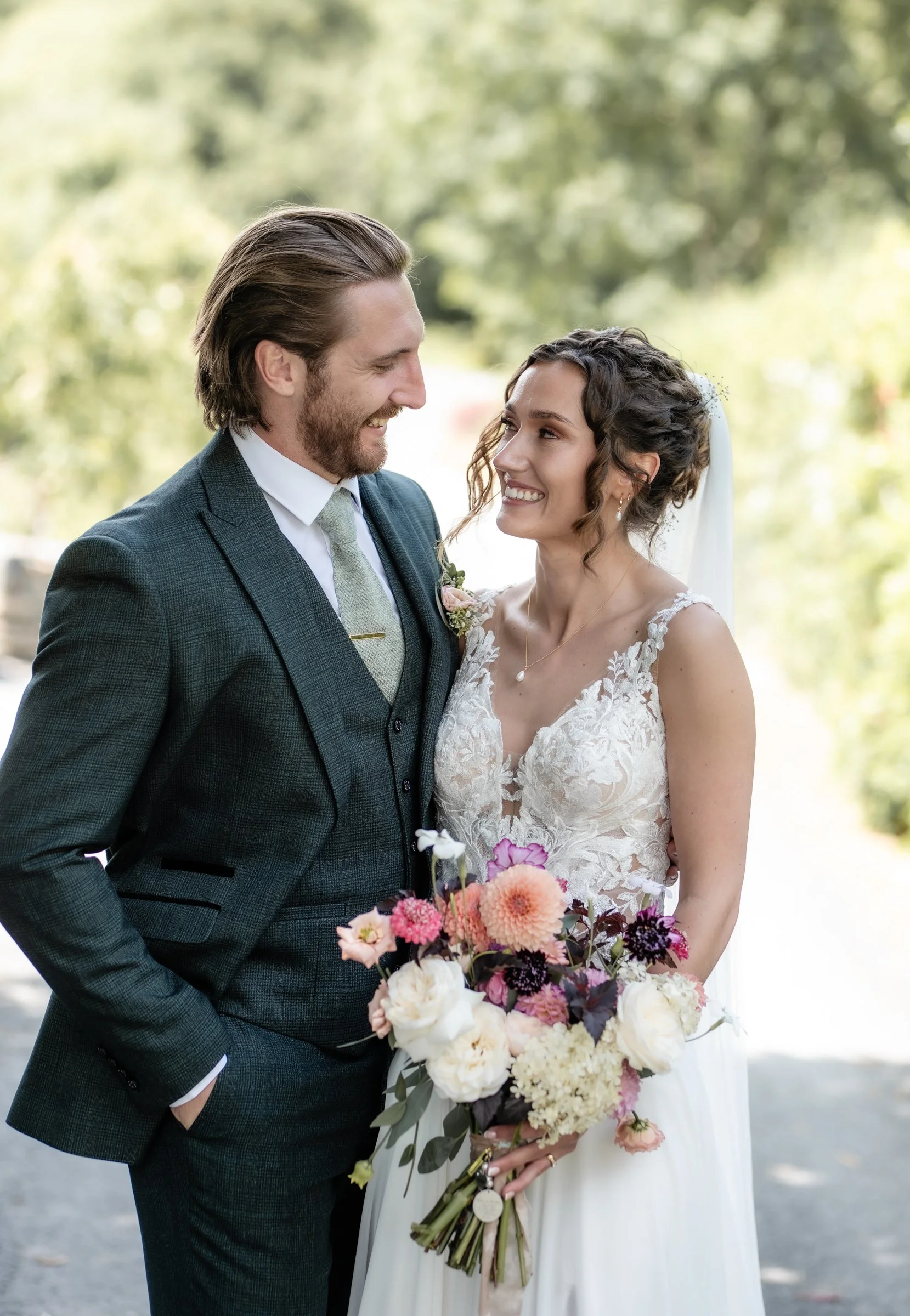 A bride and groom smiling at each other outdoors with trees in the background. The bride holds a bouquet of pink, purple, and white flowers. The groom wears a dark checked suit with a light tie, and the bride wears a white lace wedding dress with jeweled earrings and a veil.