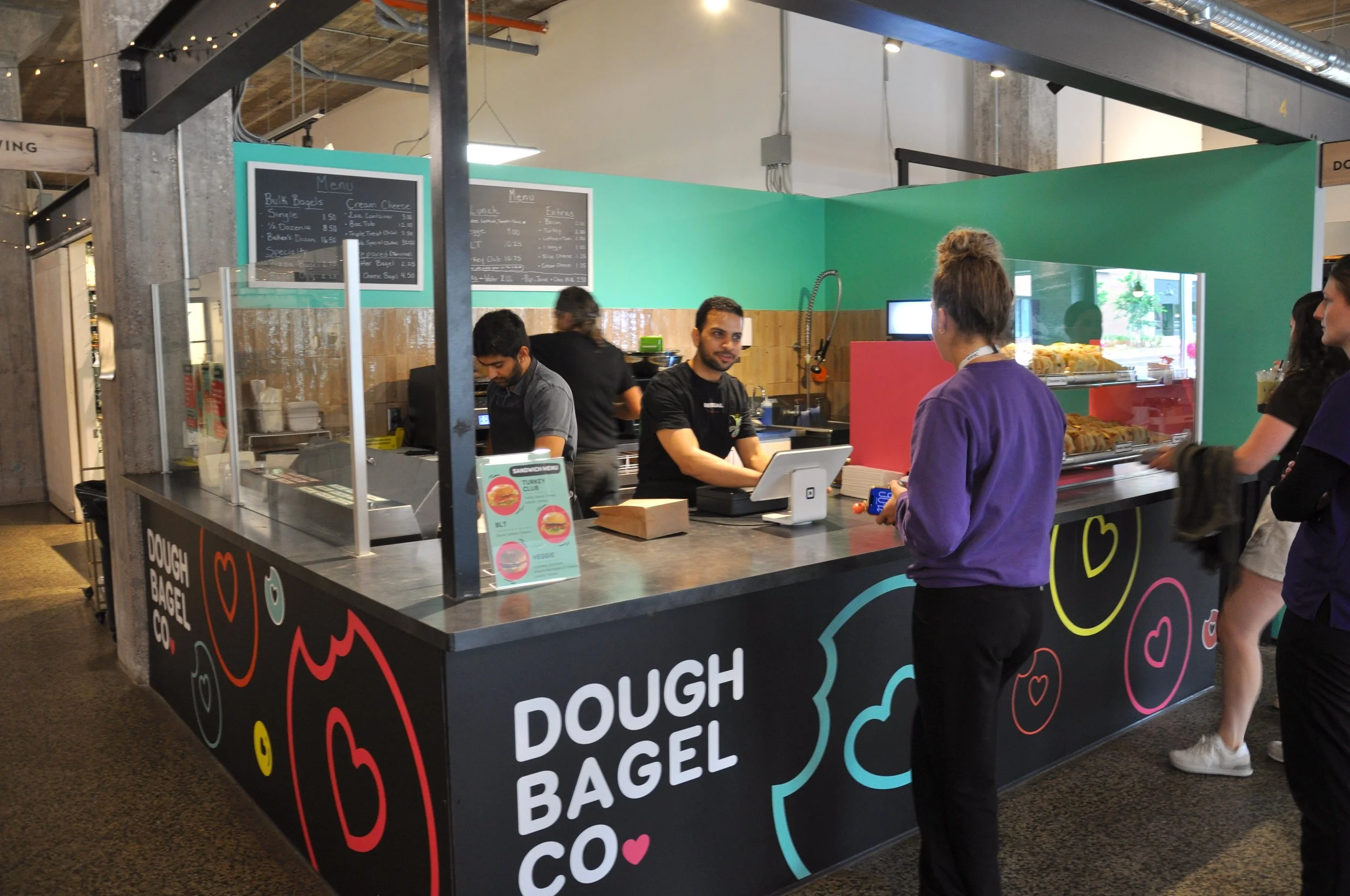 Dough Bagel Co's location in the Goods & Co. food court, featuring their colourful logo along the storefront with employees behind the counter busy preparing orders