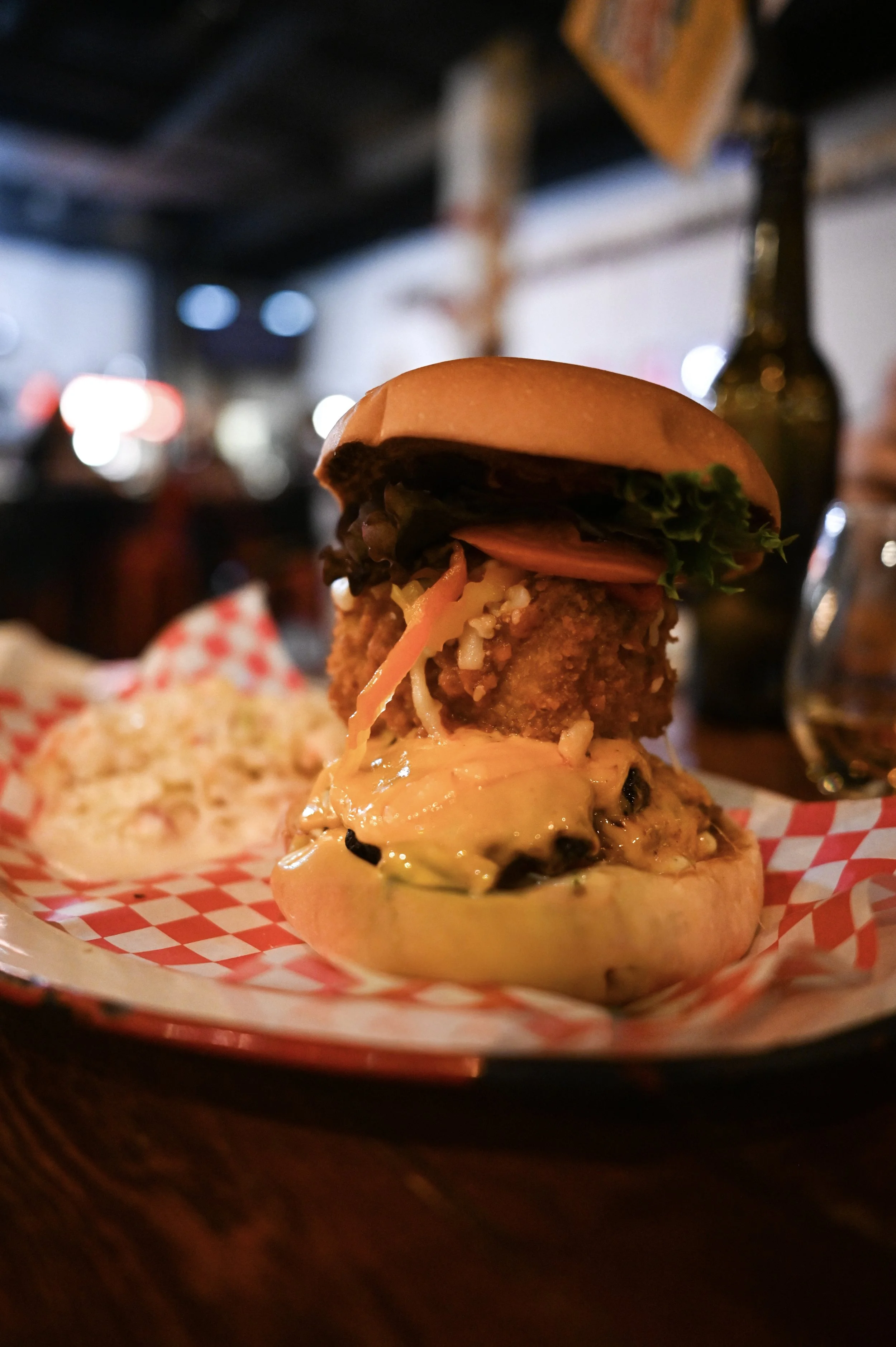 A close-up of a gourmet burger with fried chicken, lettuce, tomato, cheese, and sauce, served on a bun with a pickled onion at a restaurant.