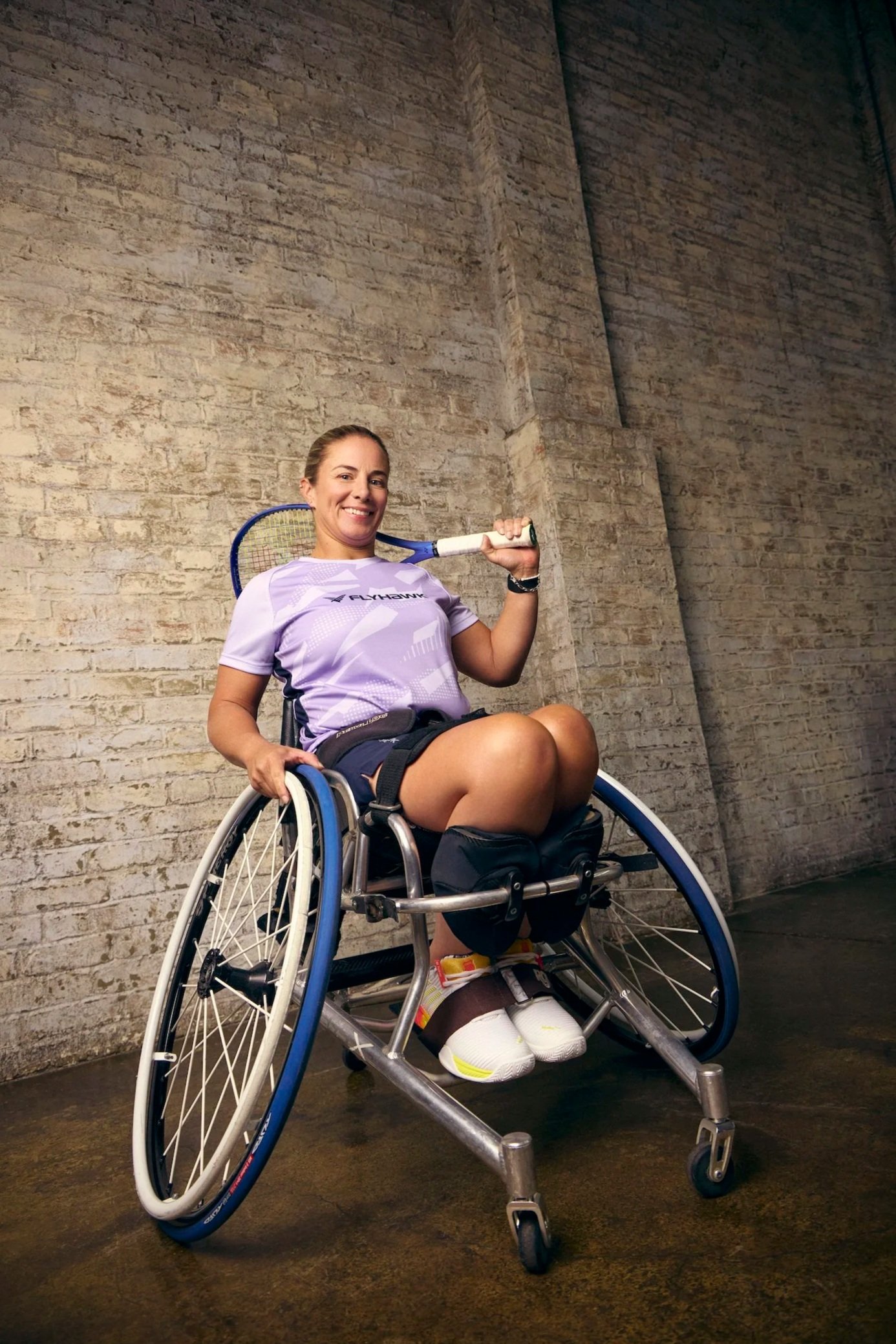 Woman in wheelchair holding tennis racket and smiling, against a brick wall.