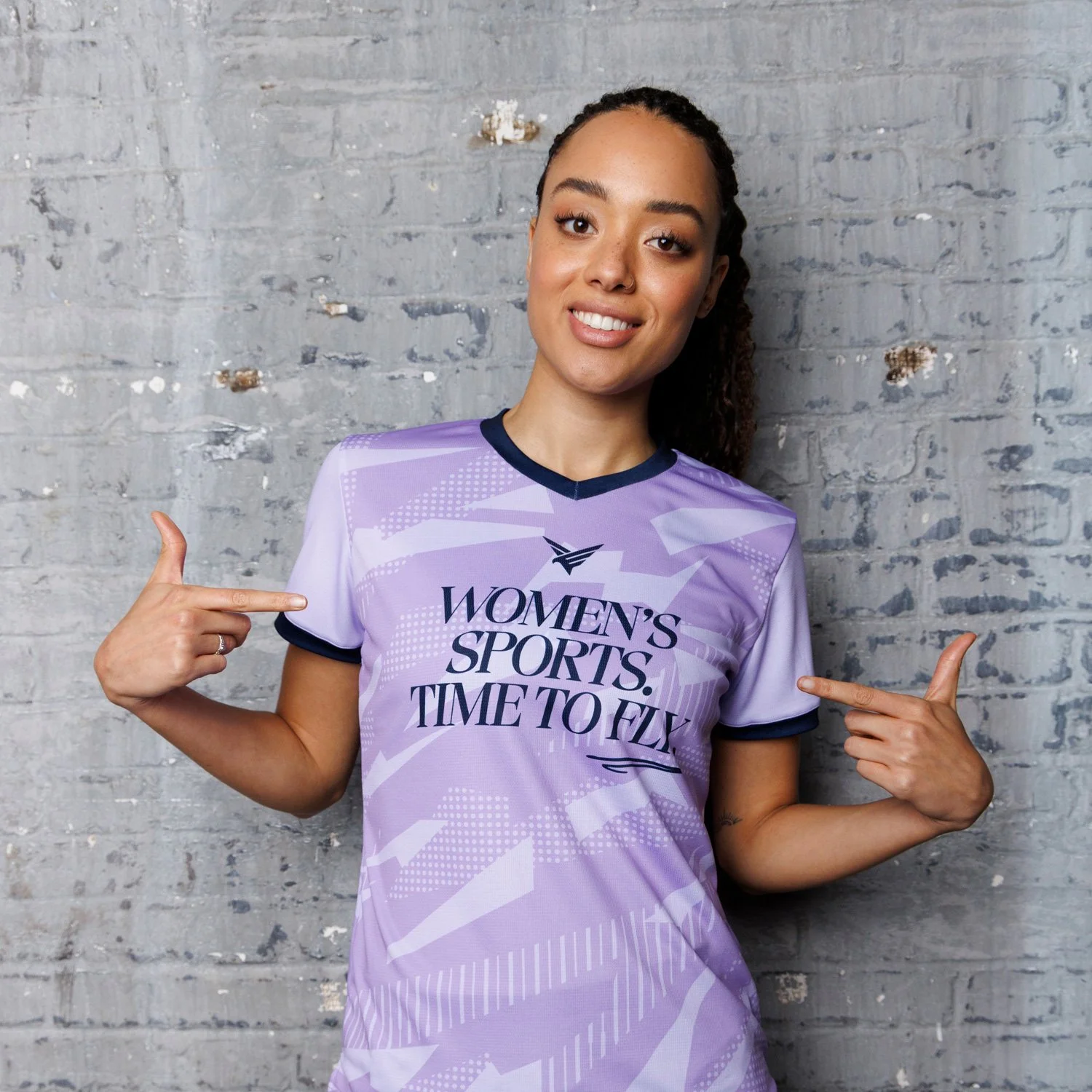 Woman in purple sports jersey pointing to the shirt with a smile against a gray brick wall.
