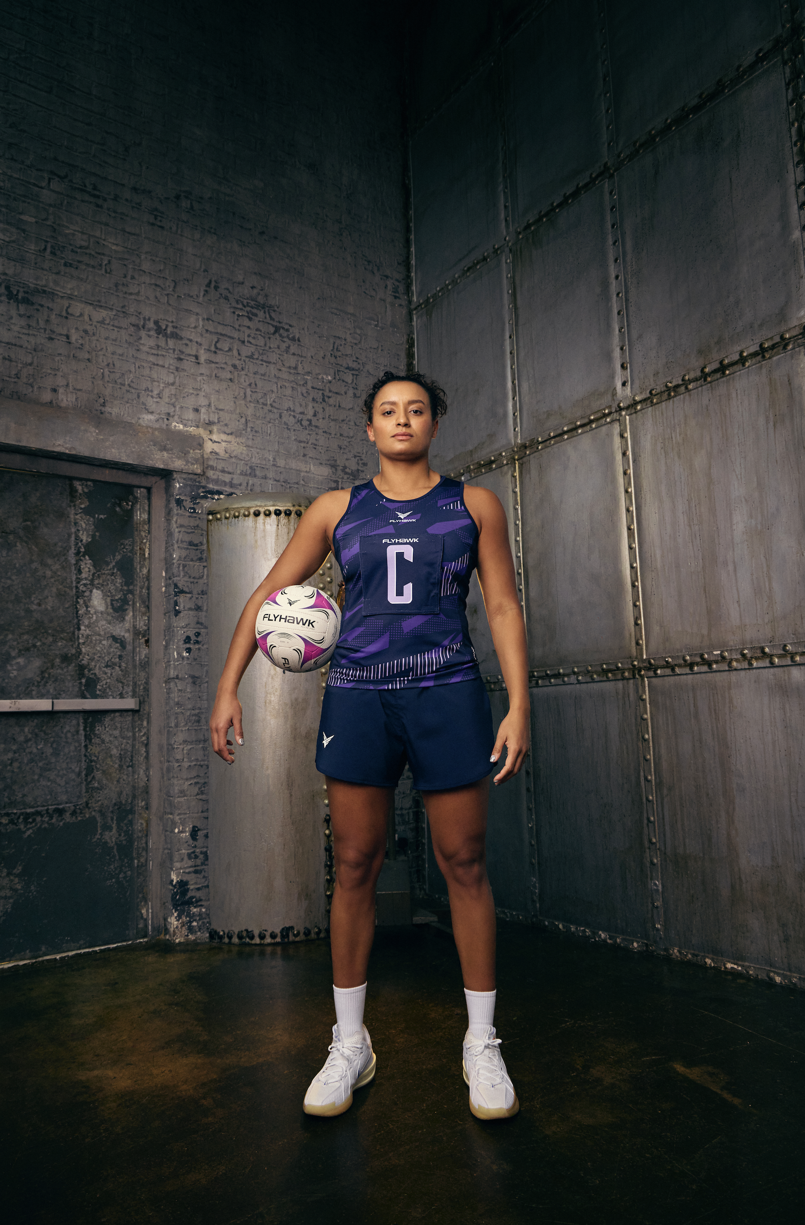 A woman in athletic gear holding a volleyball, standing in a gritty industrial-style room with metal walls and concrete floor.