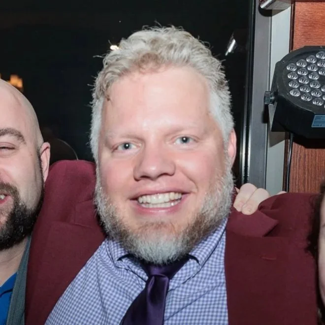 Close-up of a smiling man with blonde hair and a beard, wearing a black coat and a checkered scarf, outdoors with a blue sky in the background.