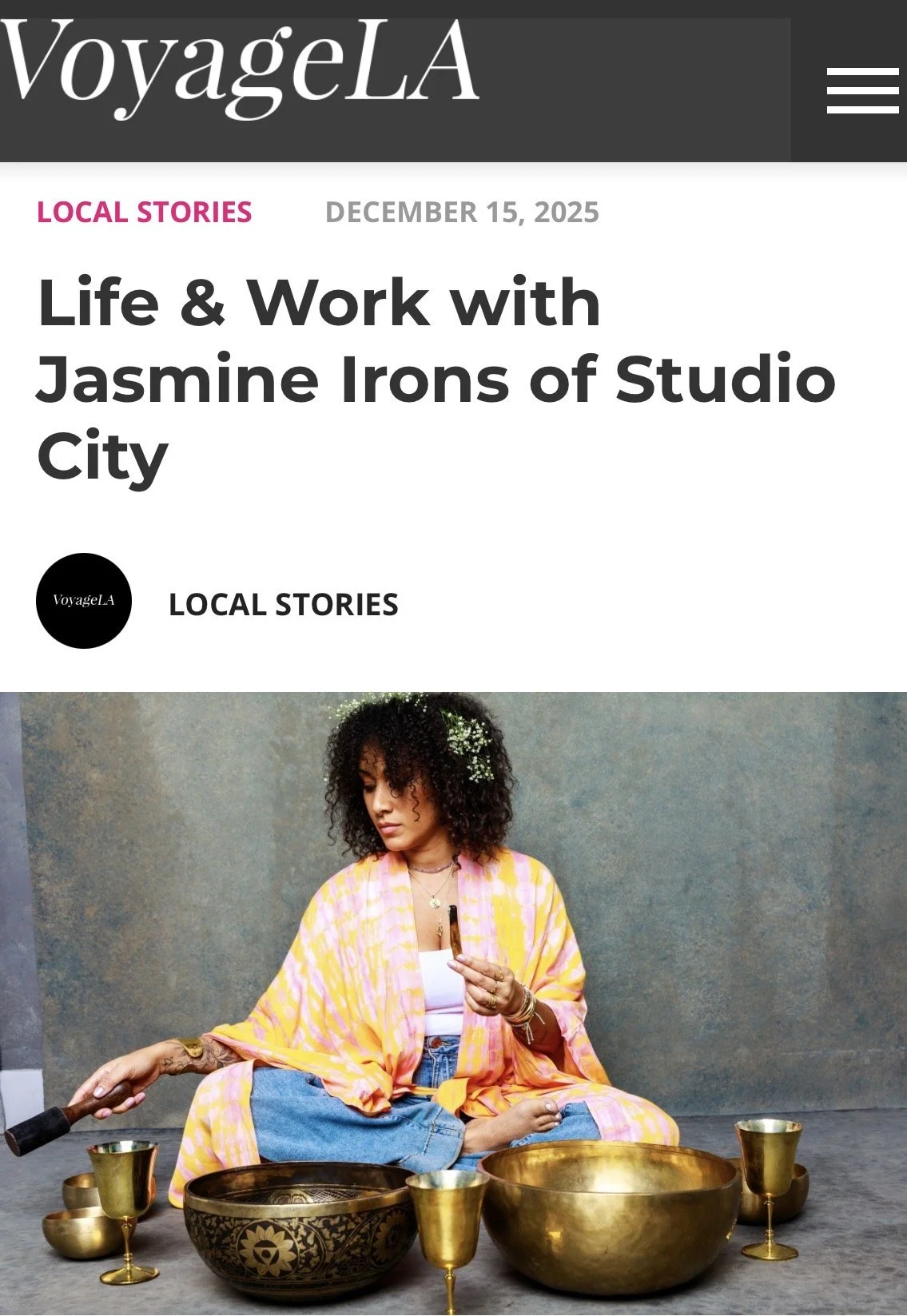 A woman sitting cross-legged on a gray surface, surrounded by singing bowls and golden chalices, holding a mallet in her right hand and a small cup in her left, with a colorful kimono and curly hair, against a muted background.
