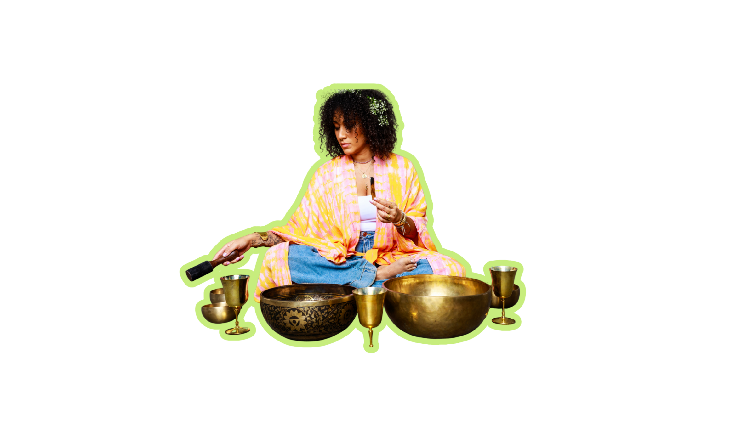 A woman with curly hair sitting cross-legged, holding a mallet, surrounded by singing bowls and cups, dressed in a colorful shawl and jeans.