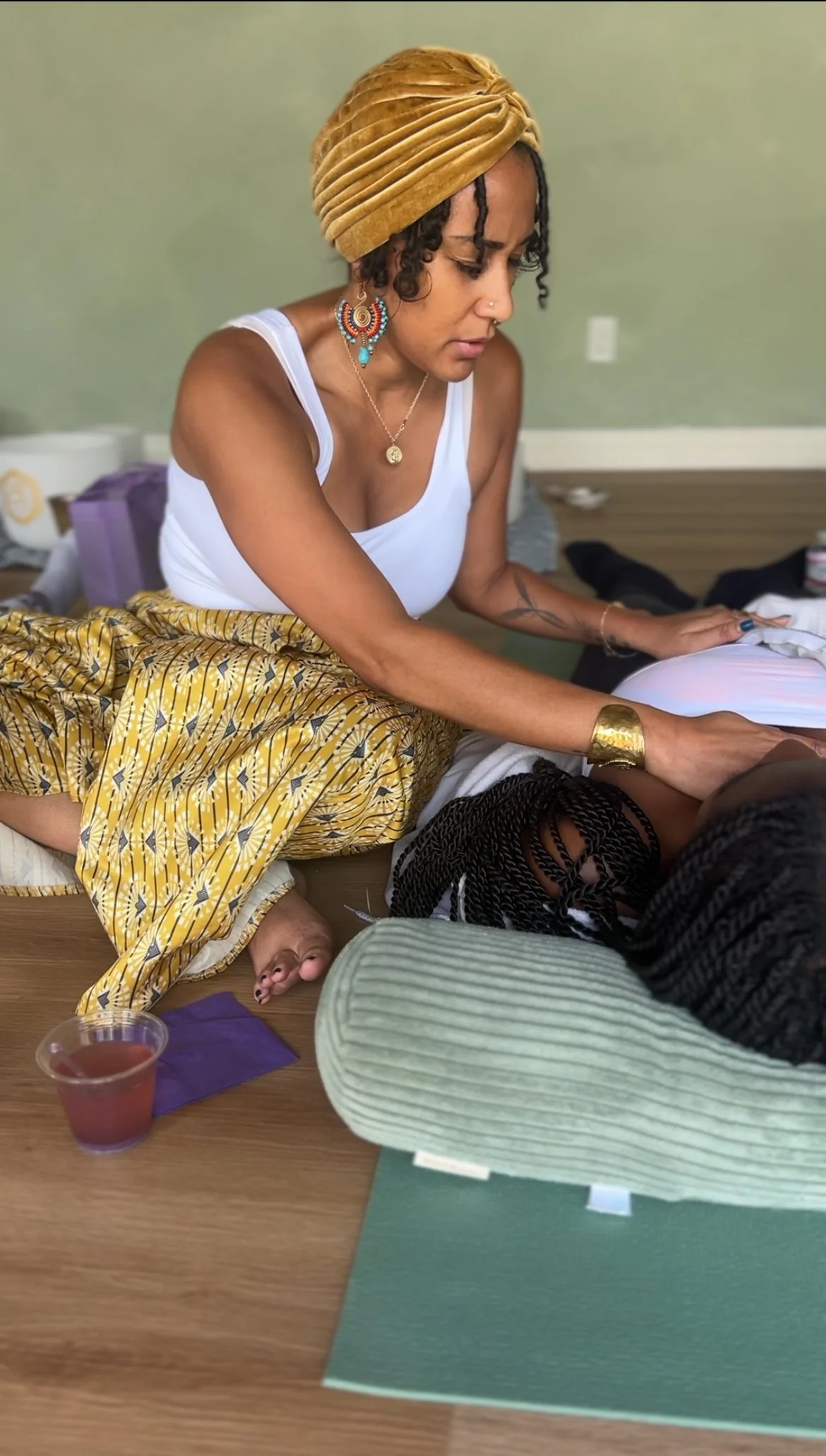 A woman with curly hair, wearing a mustard-colored turban, white tank top, and colorful jewelry, is practicing reiki on another person lying on the floor. The scene includes meditation mats, a drink in a plastic cup, and various objects in the background.