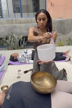 A woman performing a sound healing session outside, holding a mallet over a large crystal singing bowl while a person lies on a mat with a smaller bowl nearby.