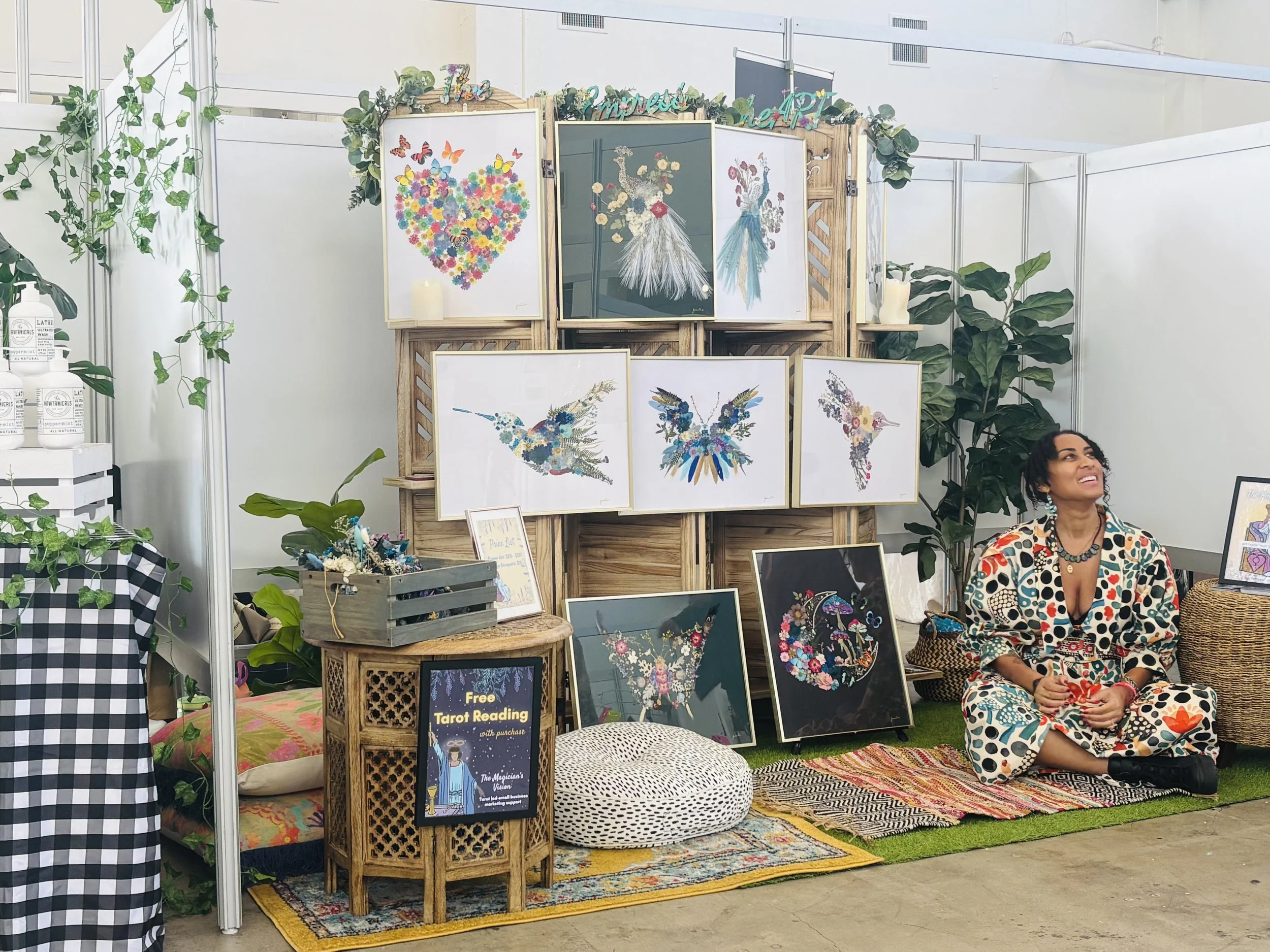 A woman sitting on the floor at an art booth, surrounded by colorful illustrations of birds and flowers, with paintings displayed on a wooden backdrop and a sign advertising free tarot readings.