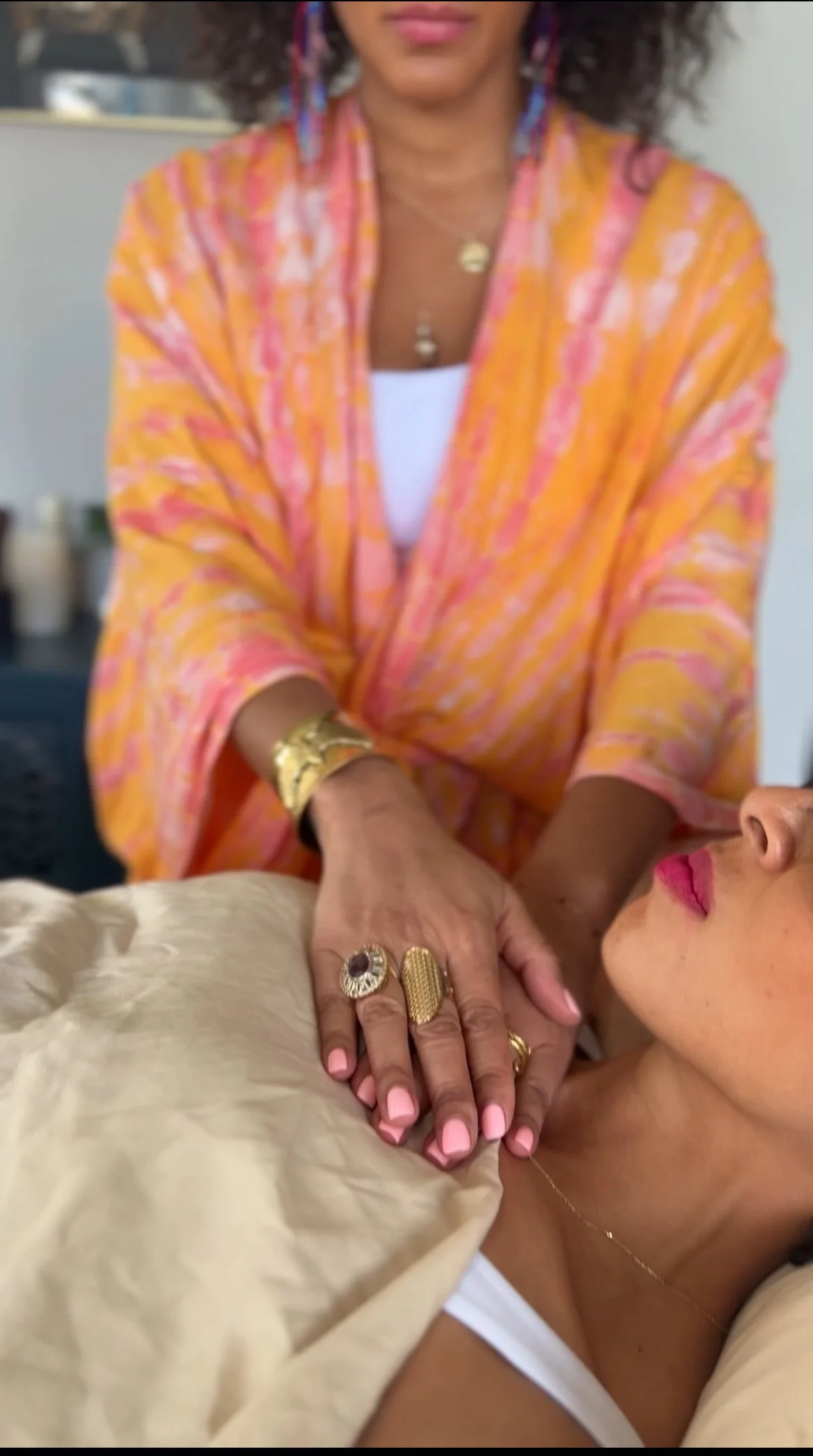 A woman receiving a massage from a therapist wearing a colorful orange and pink robe. The woman's face is partially visible, with pink lipstick and a delicate necklace. The therapist wears rings, a gold bracelet, and colorful earrings, and has her hand placed on the woman's shoulder or chest area.
