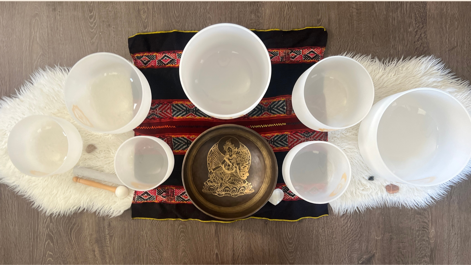 Six white bowls filled with water are arranged on a colorful embroidered cloth, with black, red, and yellow patterns. A brass tray with a decorative design featuring a deity or mythological figure and a staff with a round white head are also on the cloth. The setup is on a white furry mat on a dark wooden floor.