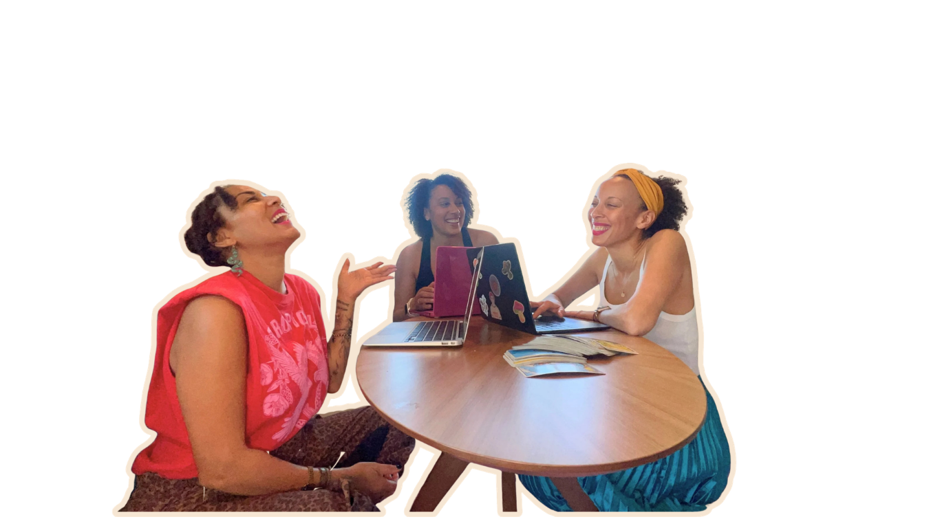 Three women sitting around a wooden table, laughing and talking, with laptops and magazines, in a cheerful meeting or discussion.