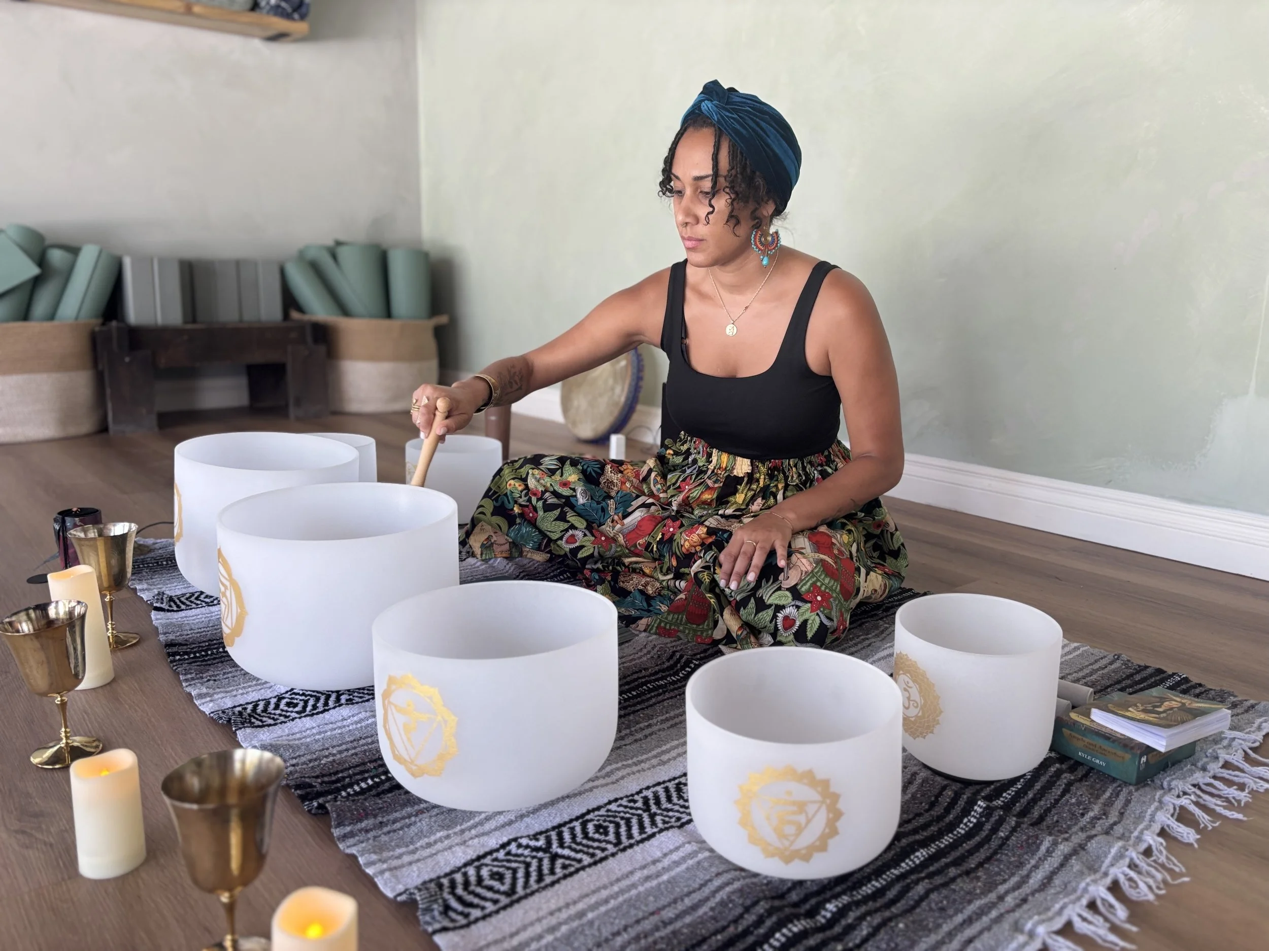 A woman with curly hair, wearing a blue headwrap and colorful earrings, is sitting on the floor playing crystal singing bowls around her. The setup includes candles, tarot cards, and various bowls on a black and white woven cloth on a wooden floor in a room with light-colored walls.