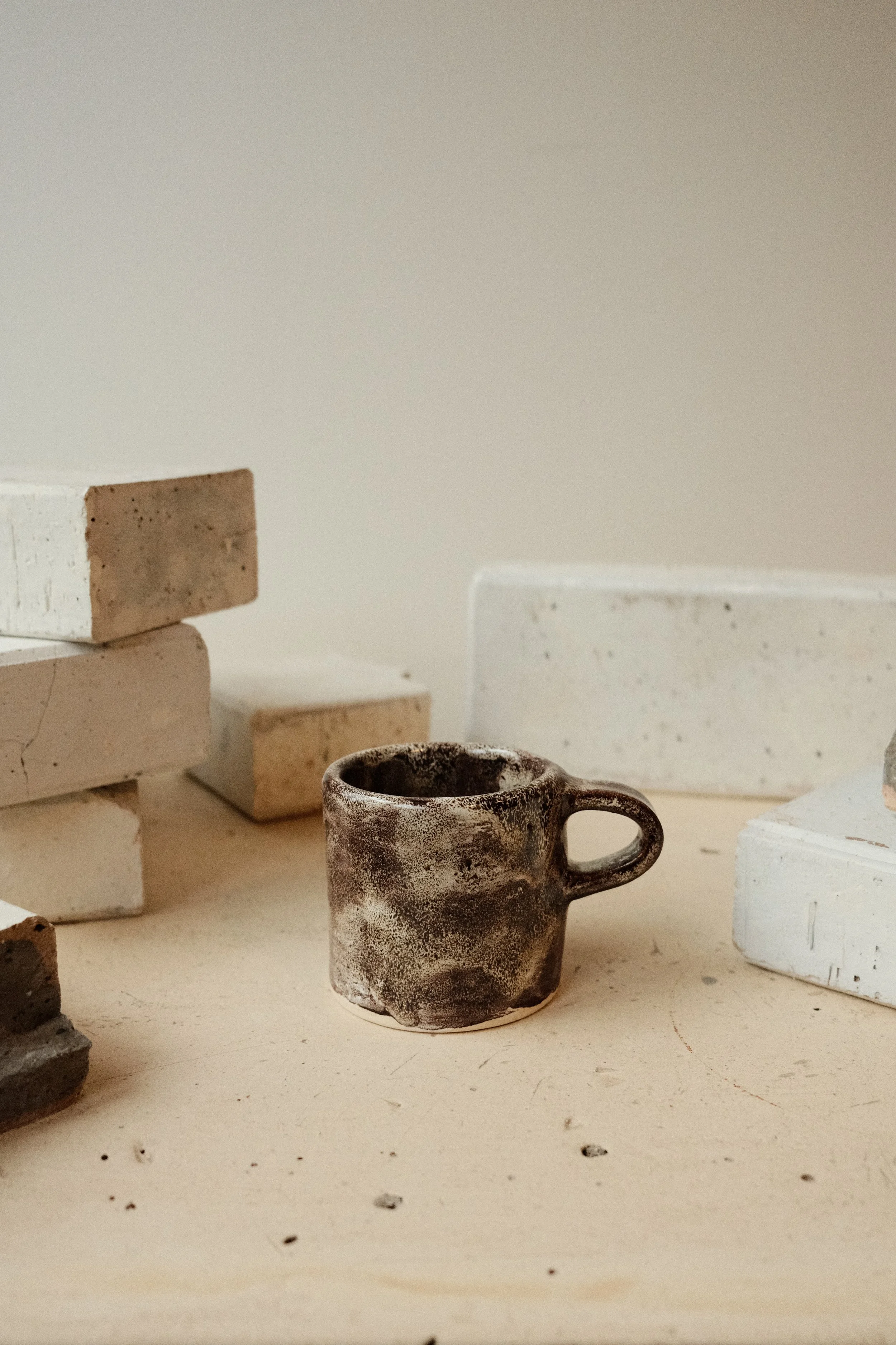 A small, rustic, dark brown ceramic mug with a handle, placed on a light-colored surface next to white and beige bricks.