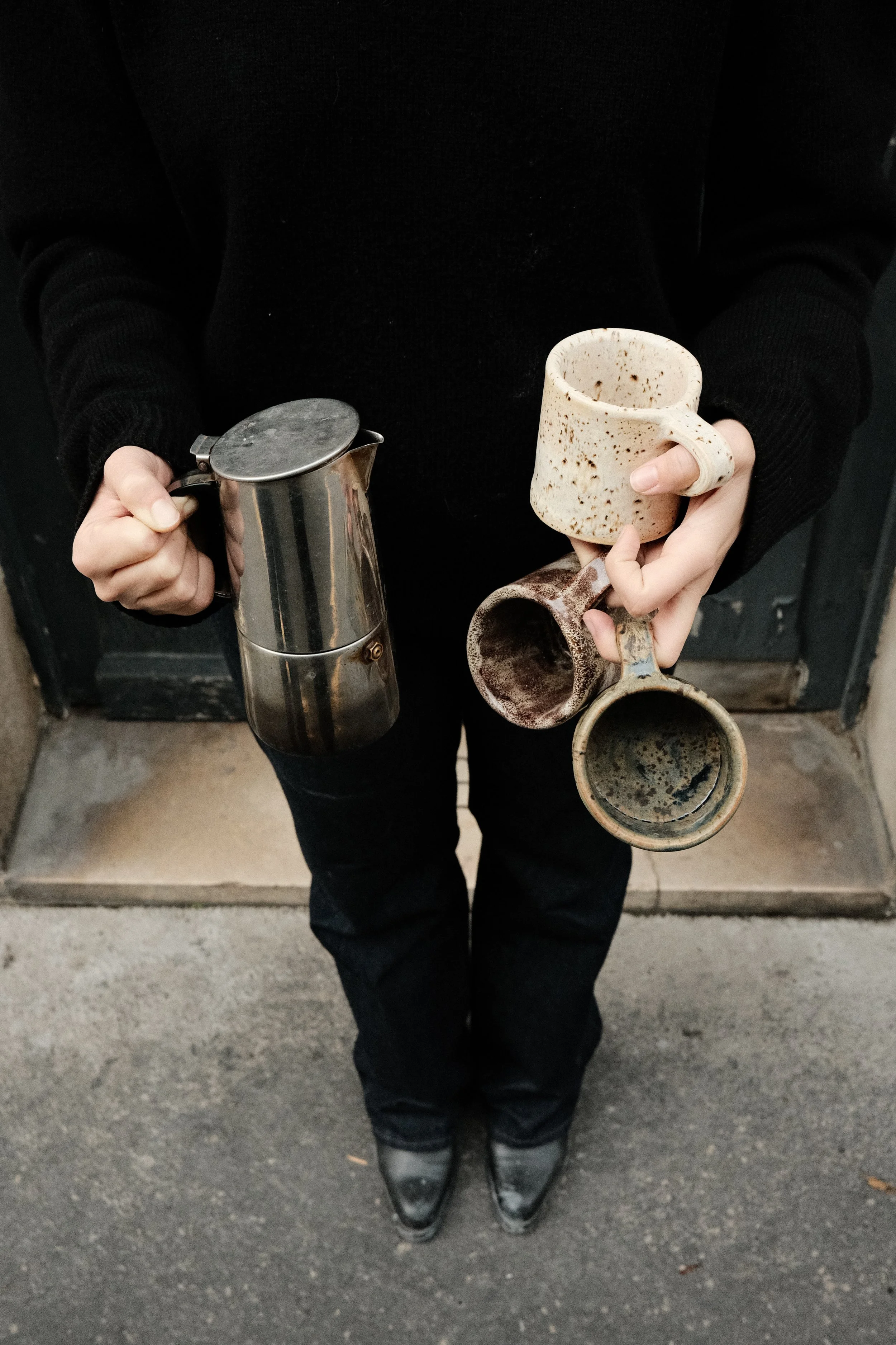 Person holding four different coffee mugs and a French press, standing on a sidewalk outside a building.