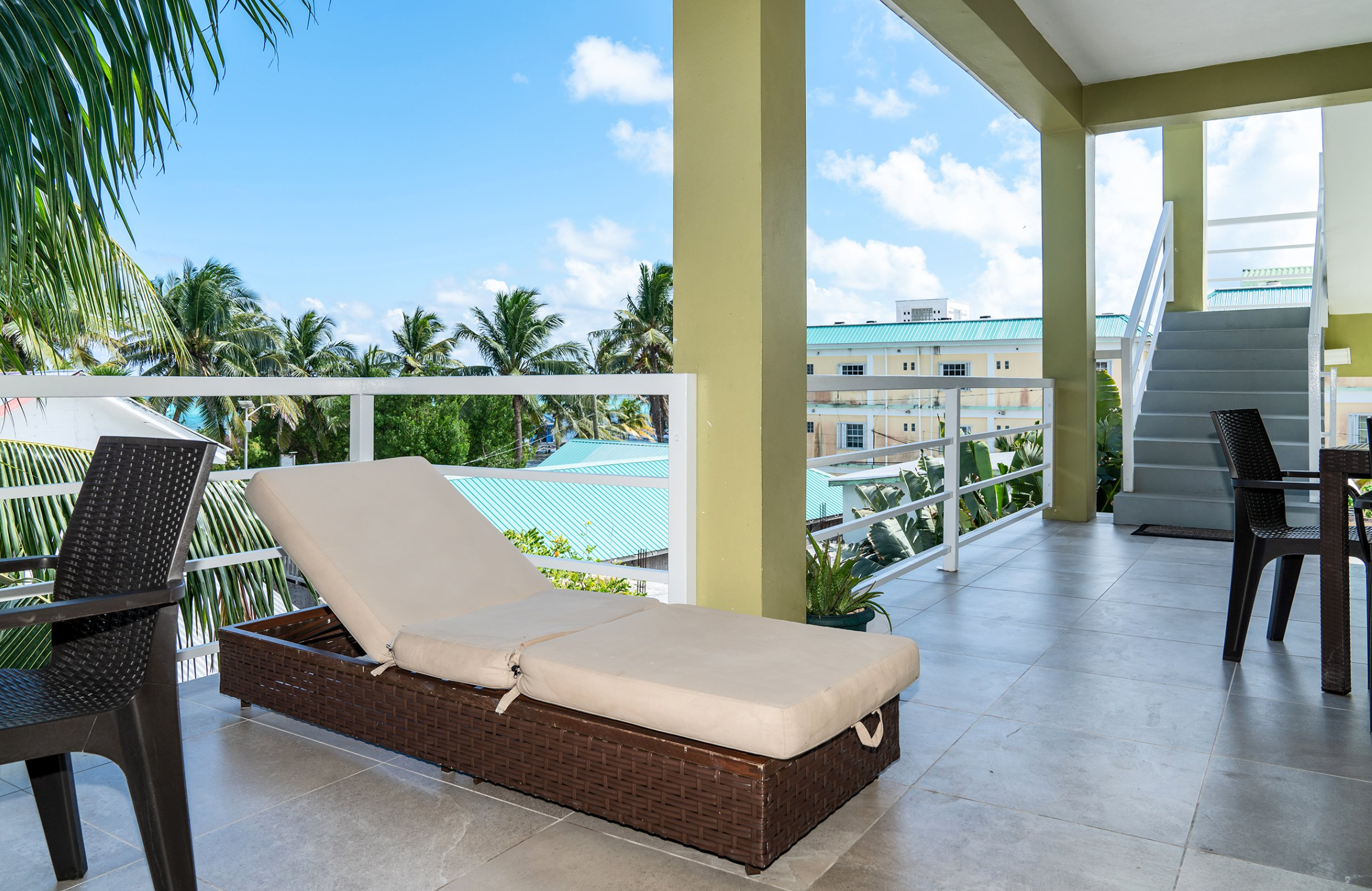 Outdoor patio with lounge chair, wicker chairs, and tile flooring overlooking palm trees and buildings with blue rooftops.