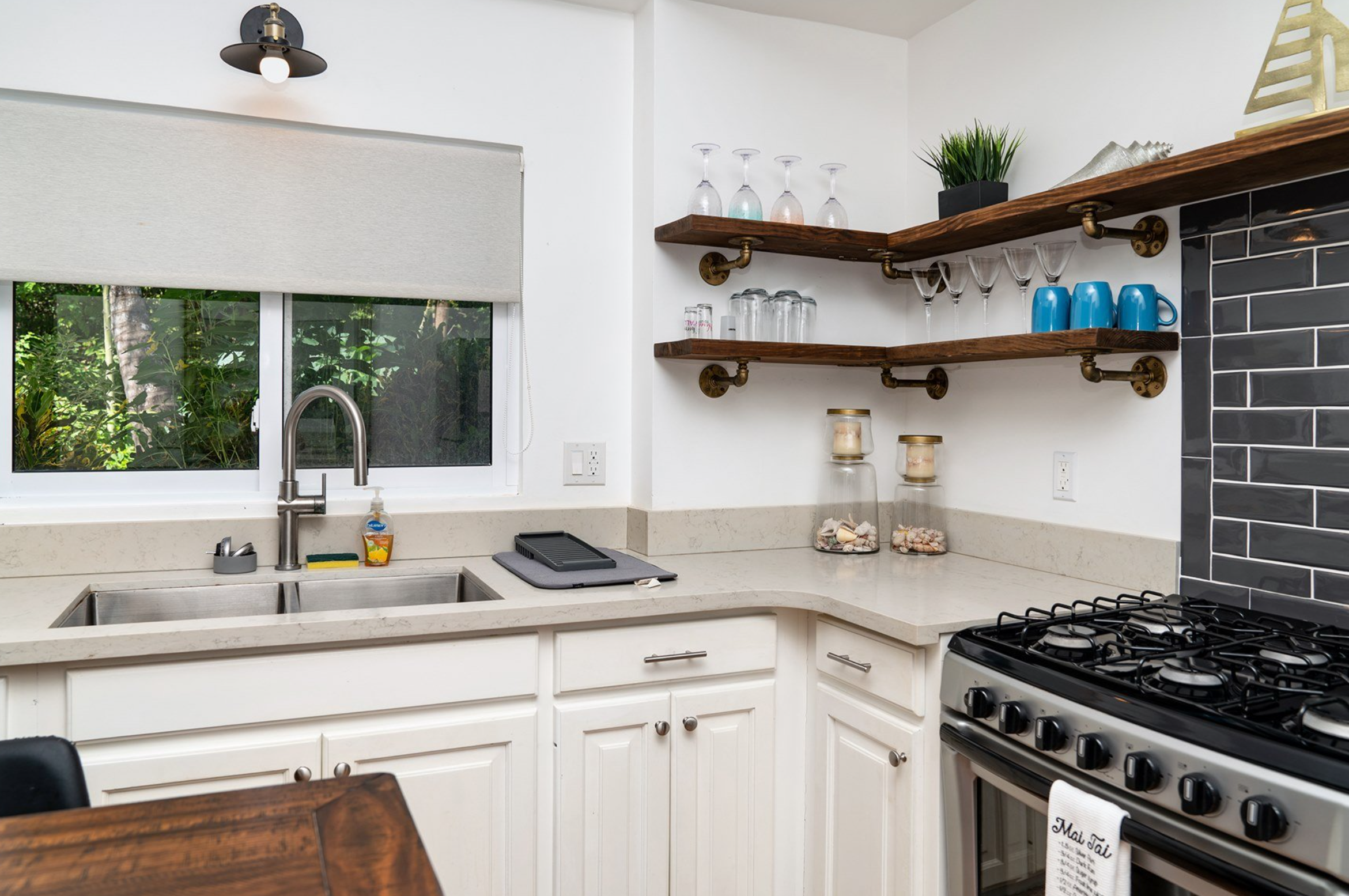 Modern kitchen interior with white cabinets, stainless steel sink, faucet, and gas stove. Open wooden shelves display glassware and blue mugs. Countertop has a tablet and soap dispenser. Window offers a view of greenery. Black tiled backsplash adds contrast.