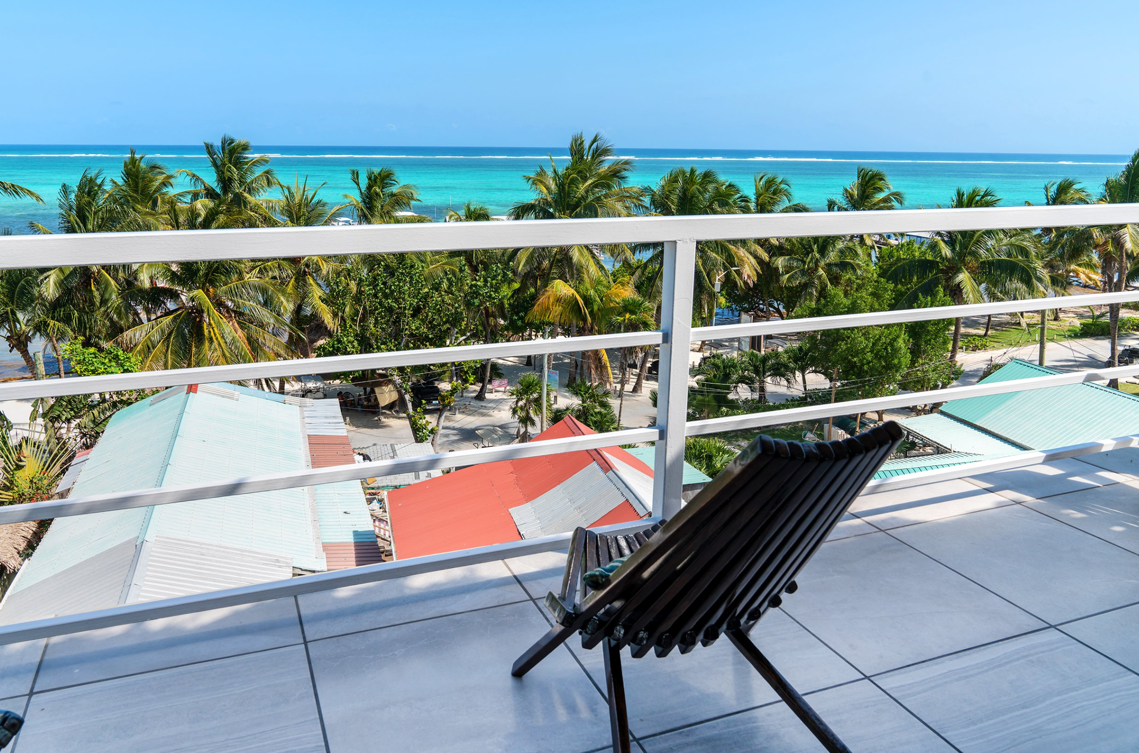 Rooftop balcony with wooden chair overlooking tropical beach, palm trees, and clear blue ocean.