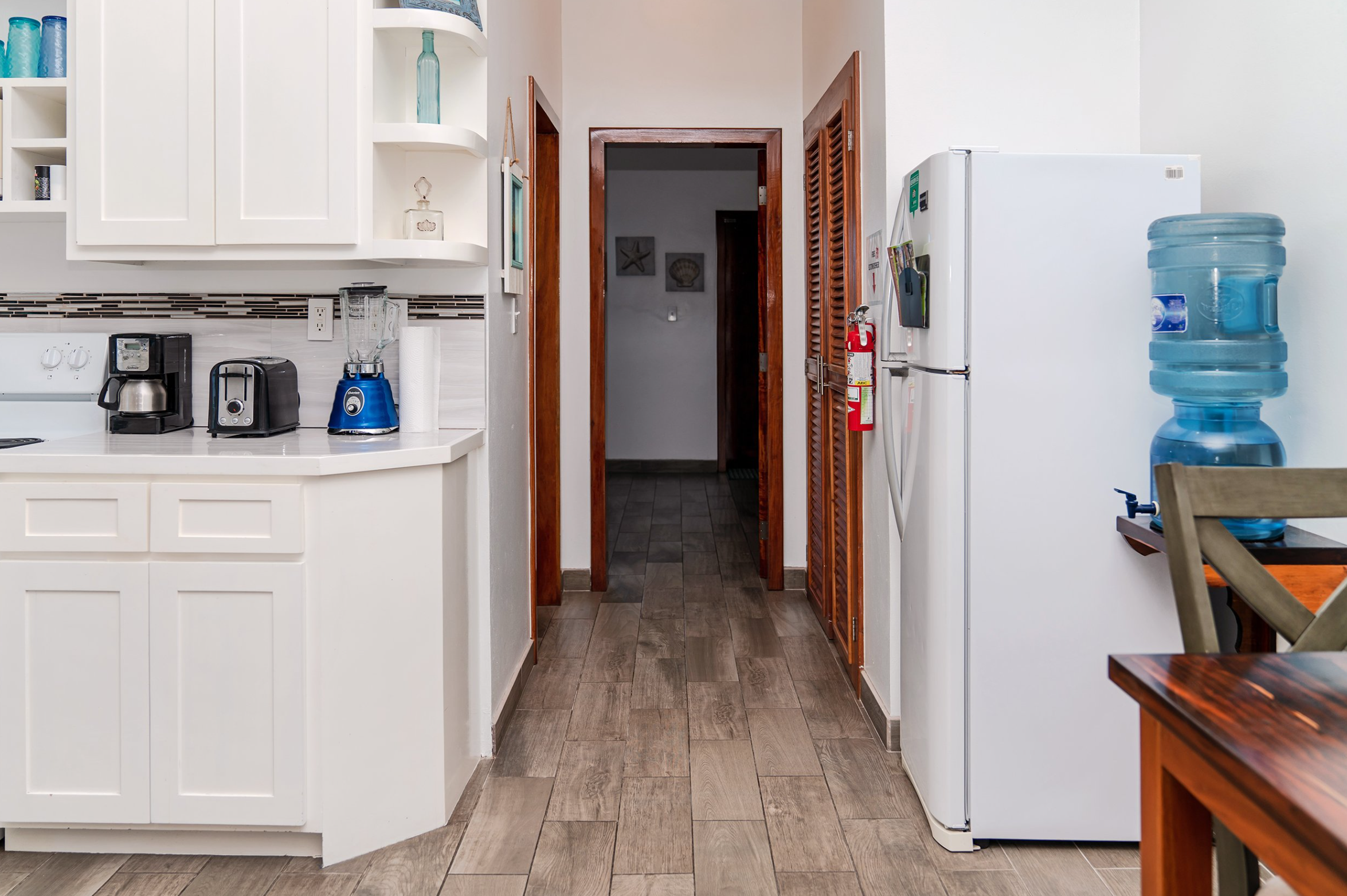 Modern kitchen with white cabinets, a refrigerator, countertop appliances including a toaster, blender, and coffee maker, a water cooler, and wooden flooring.