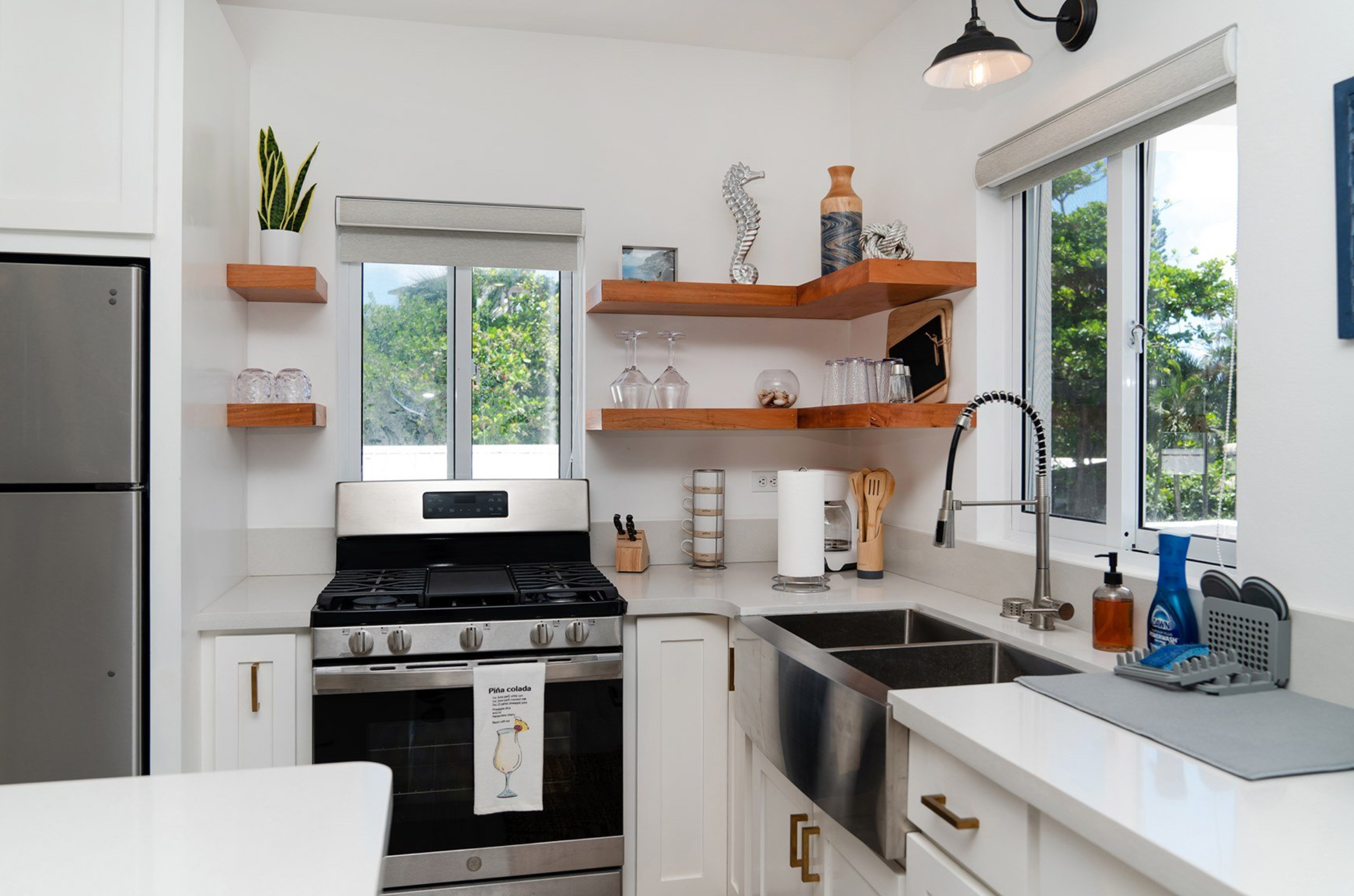 Modern kitchen with white cabinetry, stainless steel appliances, wooden shelves adorned with plants and decorative items, and a double-basin sink under a large window offering a view of greenery outside.