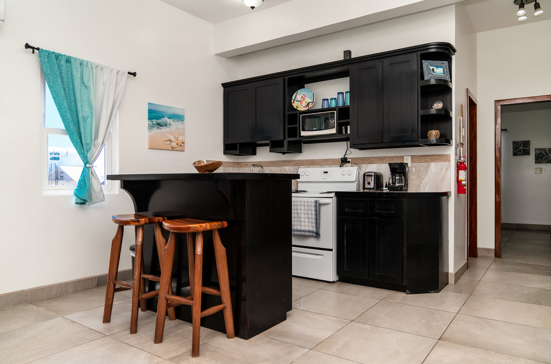 Modern kitchen with black cabinets, wooden bar stools, and appliances including a stove, microwave, and coffee maker. A turquoise curtain covers the window and a beach-themed painting is on the wall. located in walkable neighborhood on ambergris caye