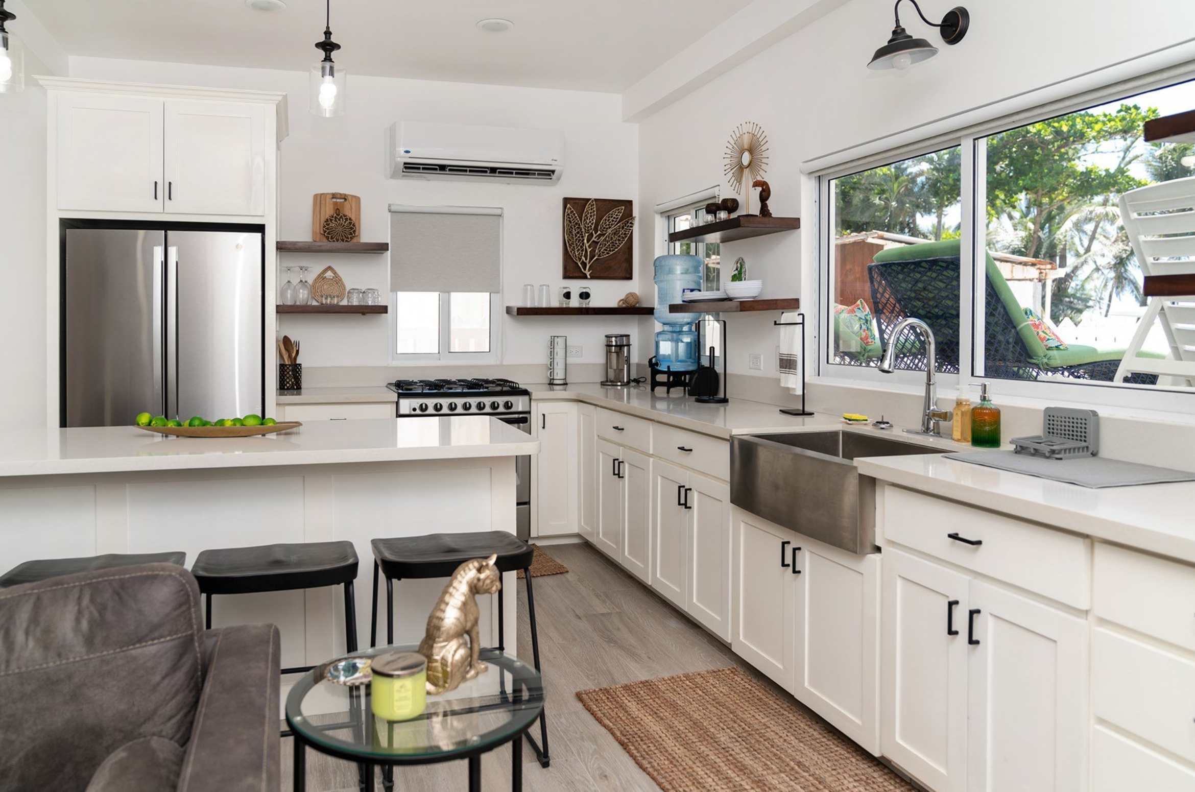 Modern kitchen with white cabinets, stainless steel appliances, breakfast bar, and decor items. Large window reveals outdoor greenery. Wood floor and two stools are visible. Belize real estate property