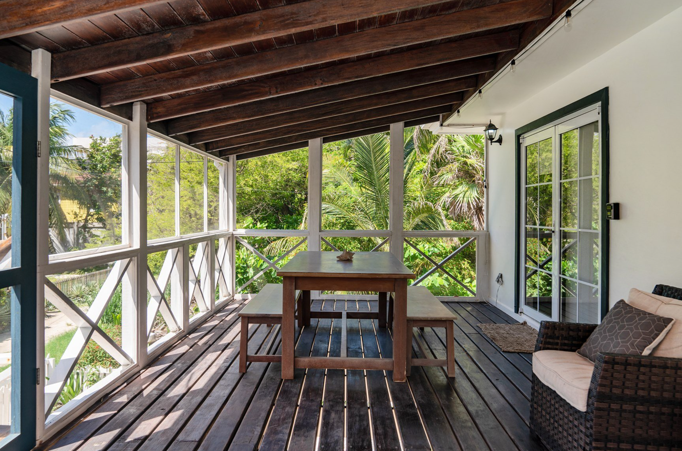Screened-in porch with wooden table, benches, wicker chair, and view of lush greenery outside.