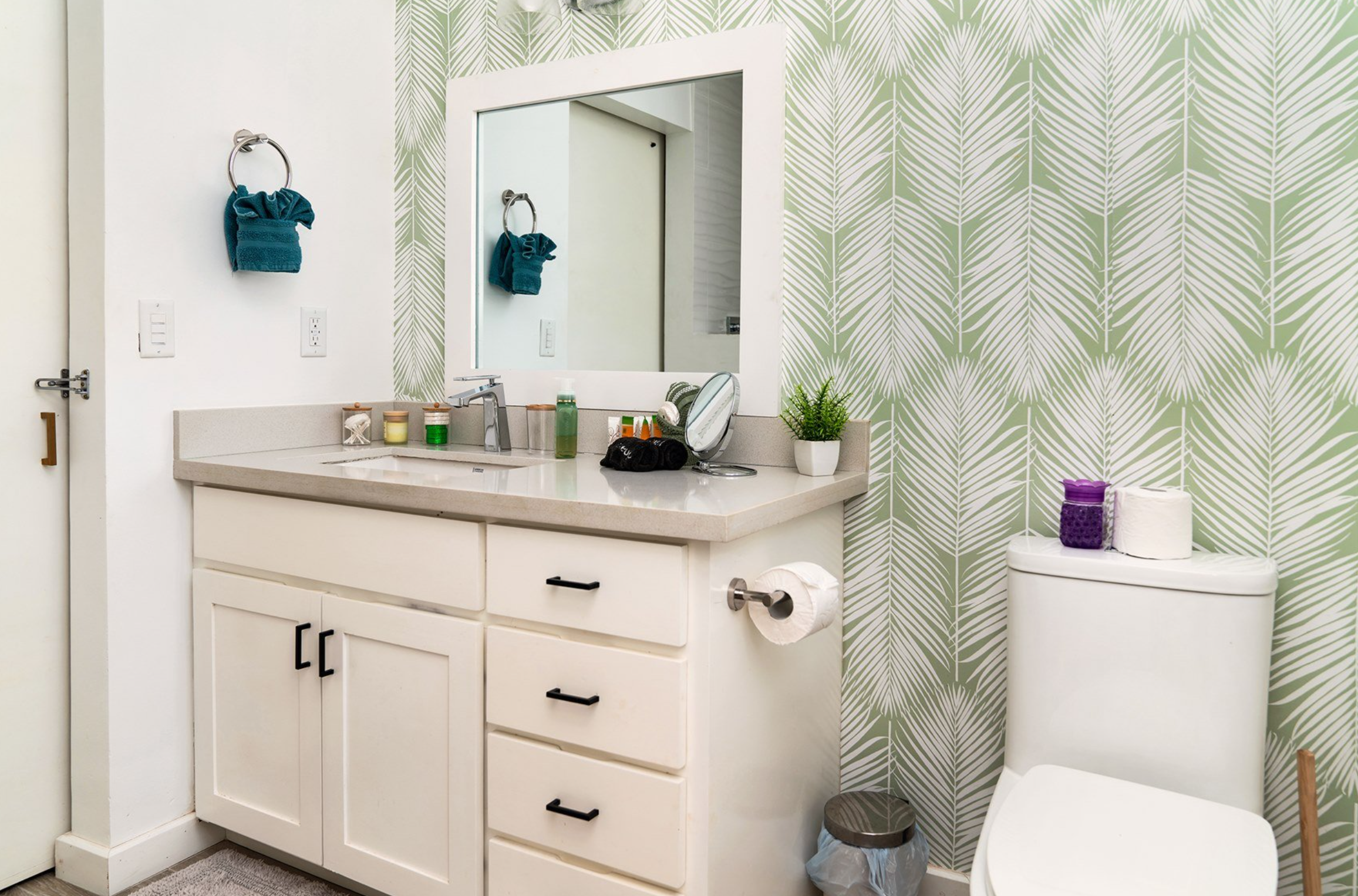 Modern bathroom with white cabinets, a countertop sink, large mirror, toilet with a purple jar and toilet paper, plants, green patterned wallpaper, and teal towels.