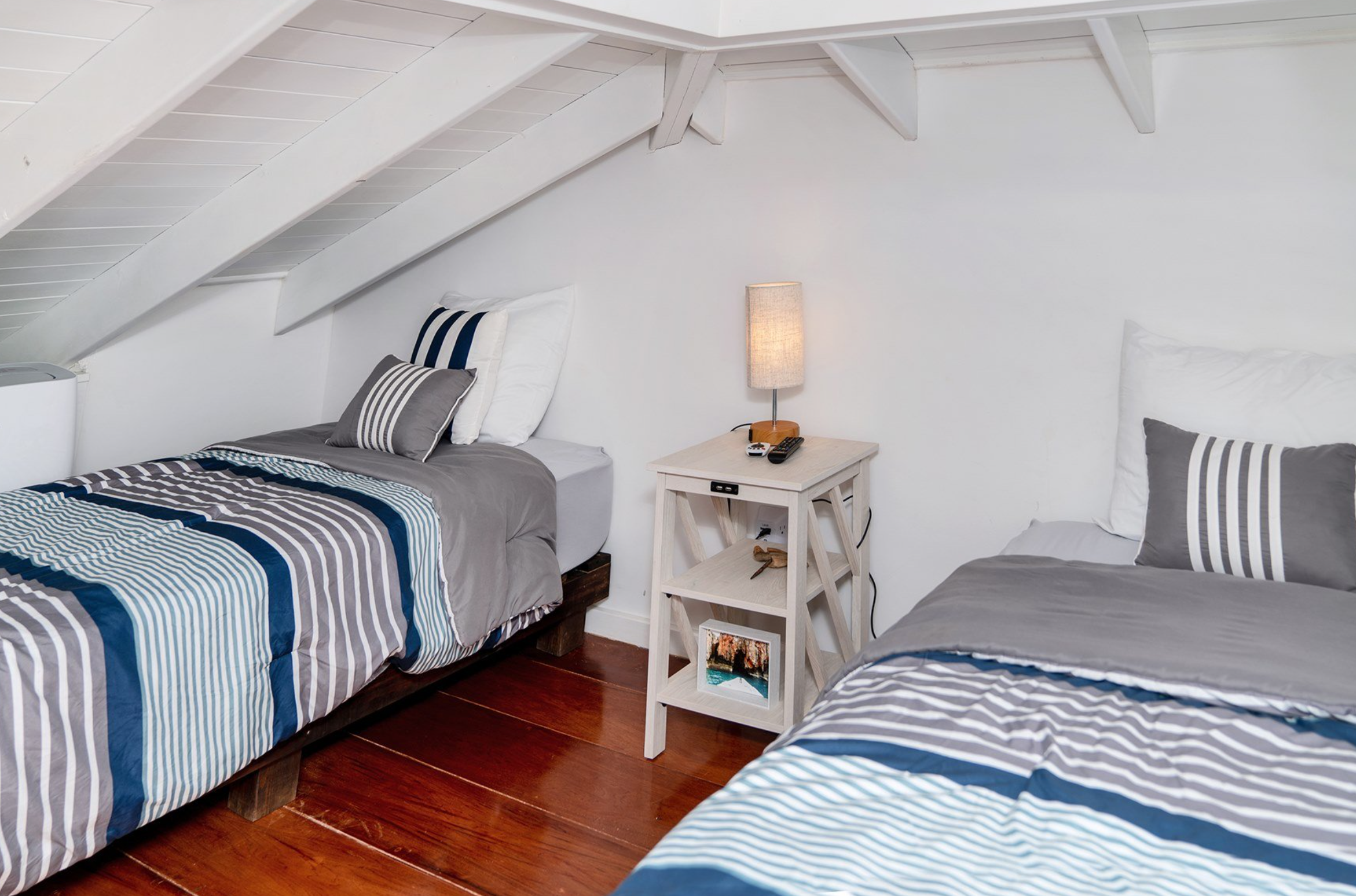 Attic room with two twin beds, striped bedding, a nightstand with a lamp, and wooden floor.