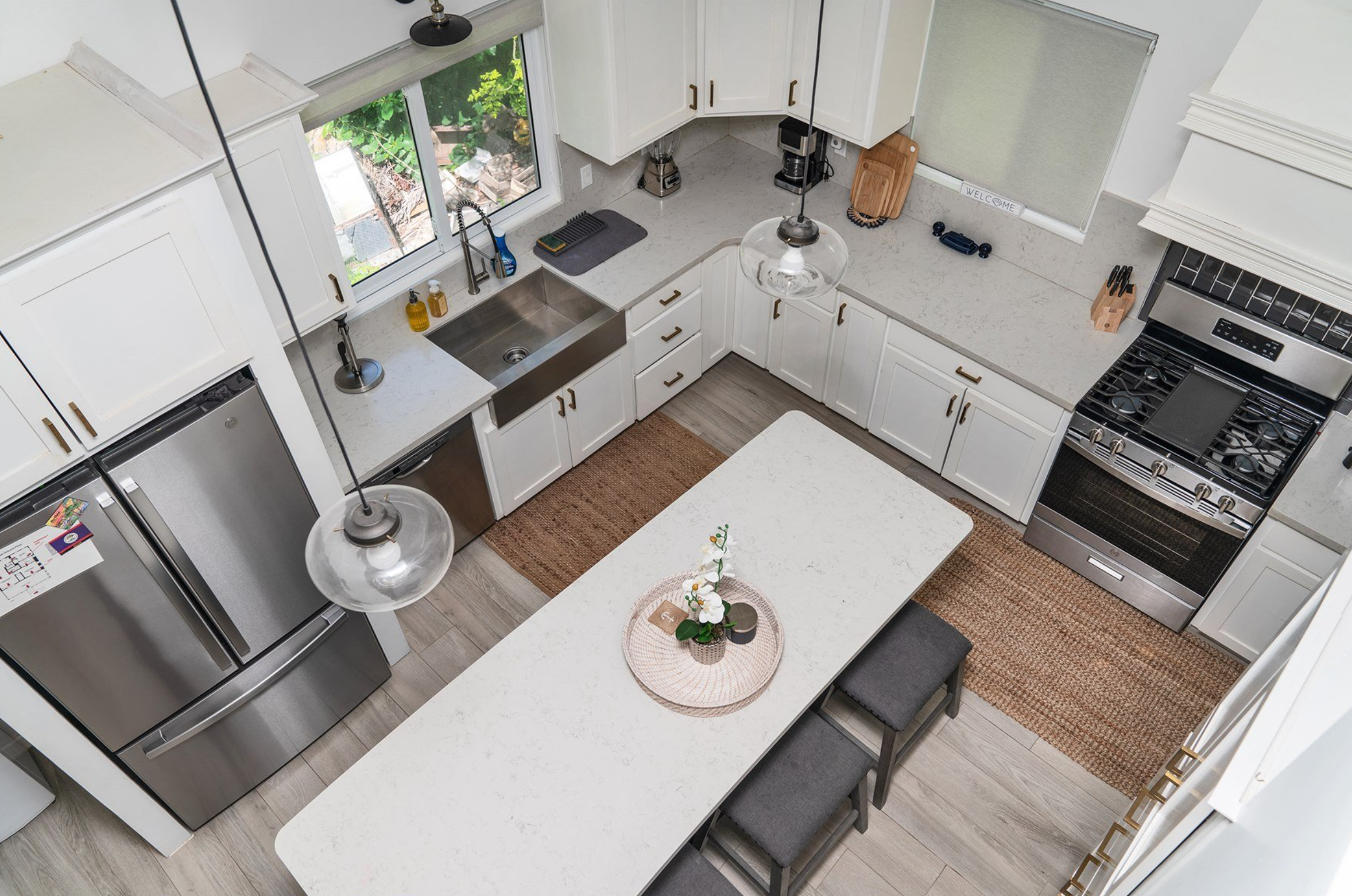 Overhead view of a modern kitchen with white cabinets, stainless steel appliances, and a central island. The kitchen features a large fridge, gas stove, dual sinks, pendant lighting, and a woven rug. A decorative tray with plants is on the island.