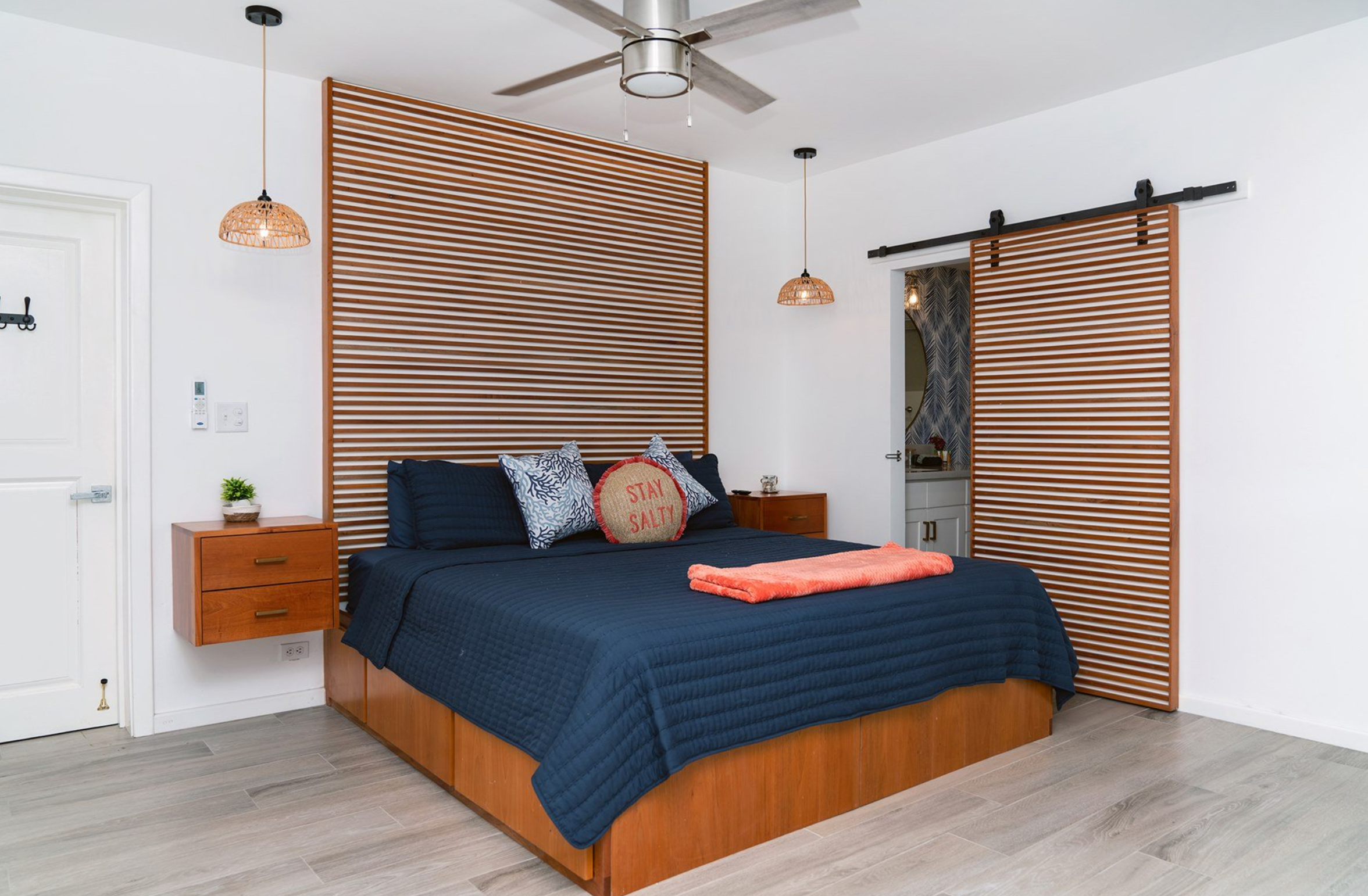 Modern bedroom with striped wooden paneling, navy bedding, and orange accents. Features a ceiling fan, pendant lights, and a sliding barn door. Bedside drawer with small plant decoration.